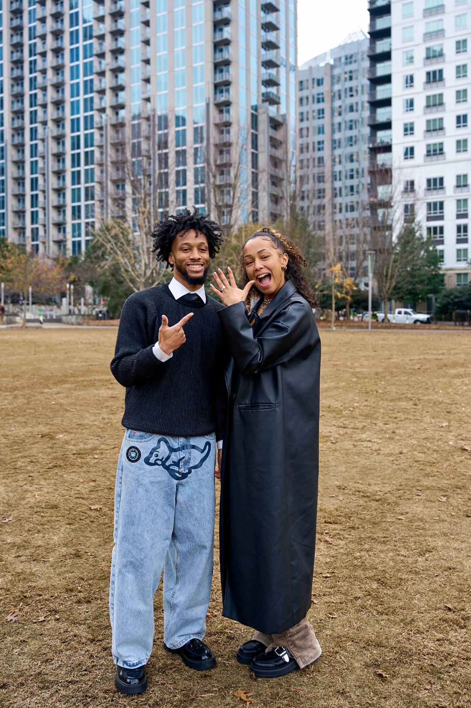 Two people pose playfully in front of modern apartment buildings on a brown grass field. North Carolina Wedding Photographer.
