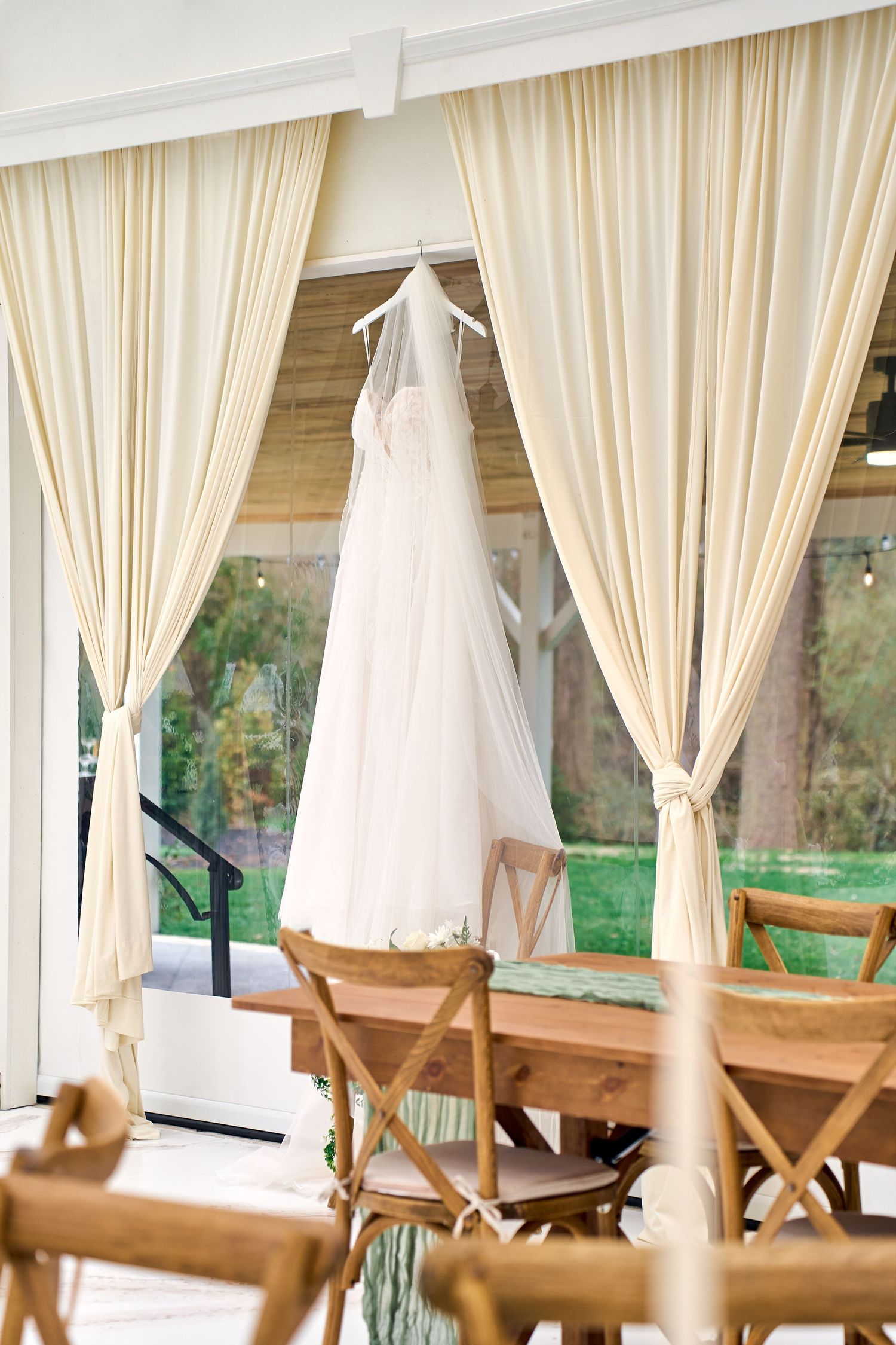 A white wedding dress hangs elegantly between cream-colored curtains in a reception area with wooden rustic chairs.