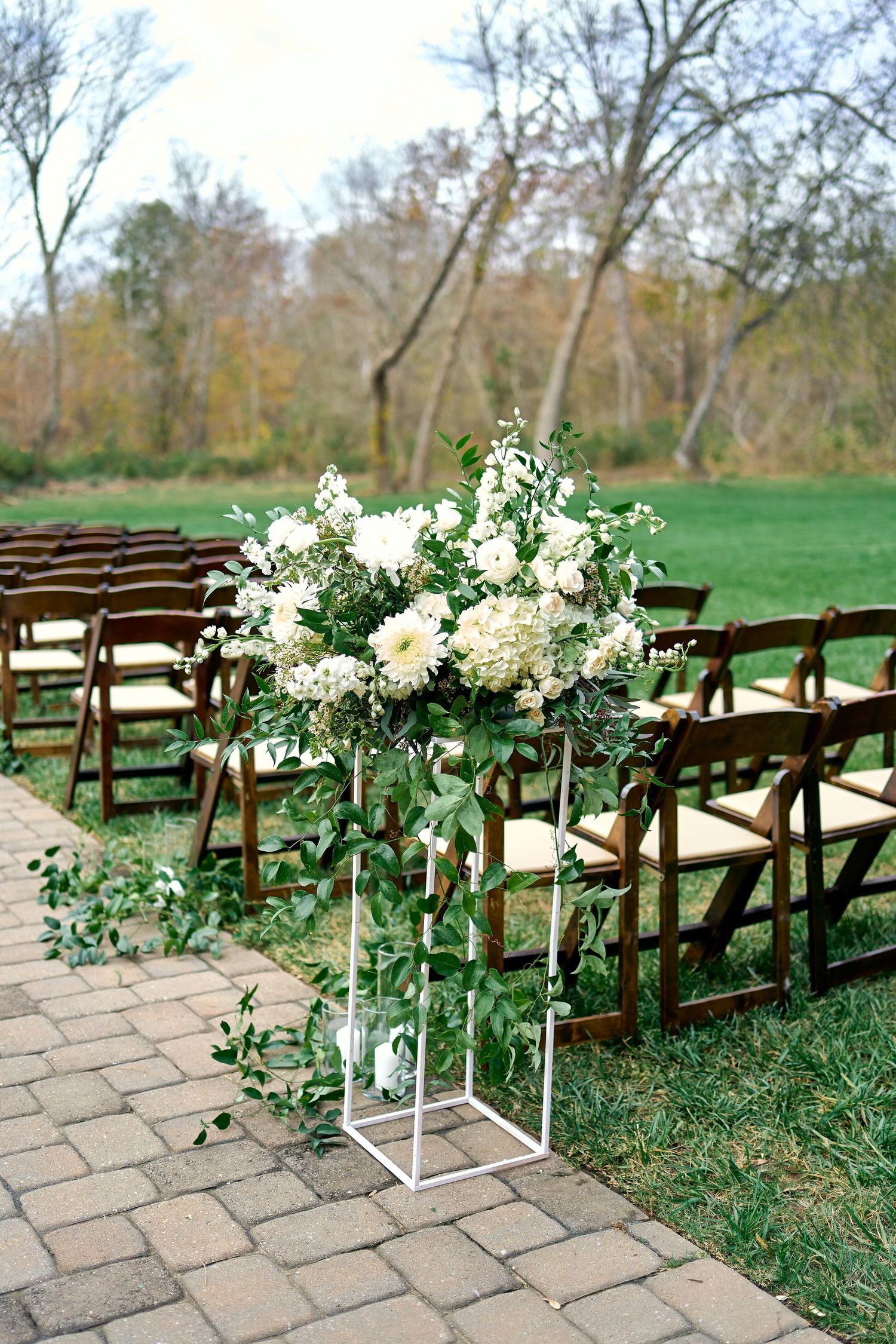 Elegant white floral arrangement on a metal stand decorates the aisle of an outdoor wedding ceremony with wooden chairs.
