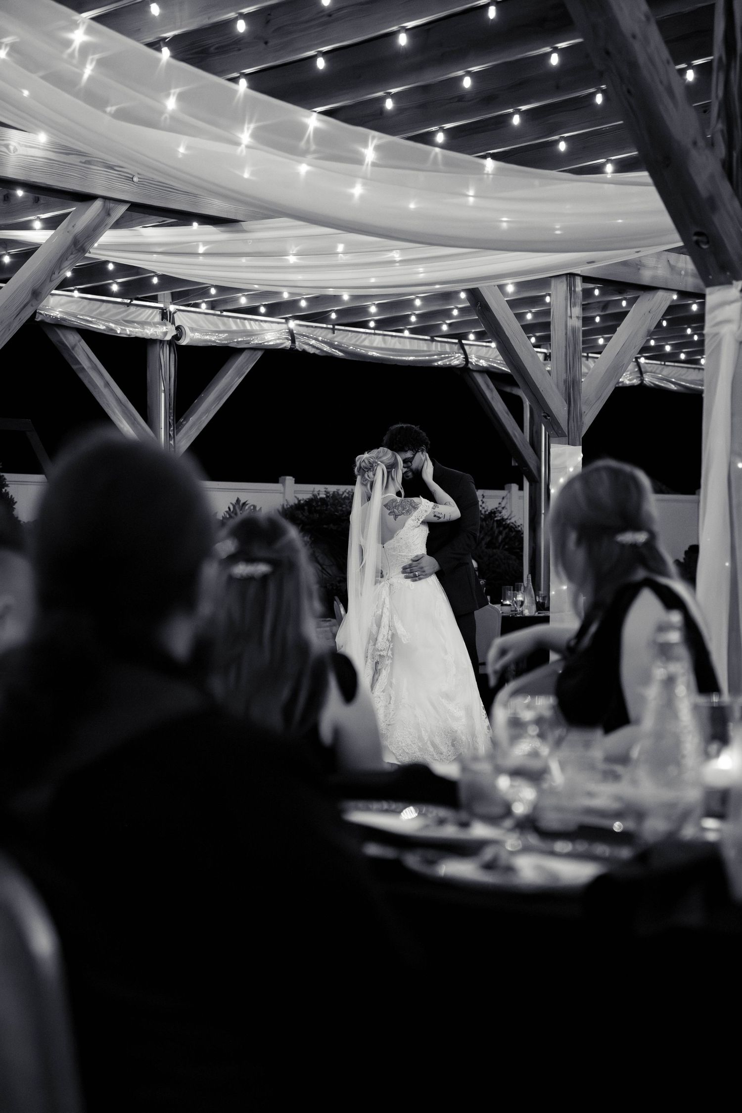 Black and white photo of bride and groom sharing first dance under string lights at outdoor reception.