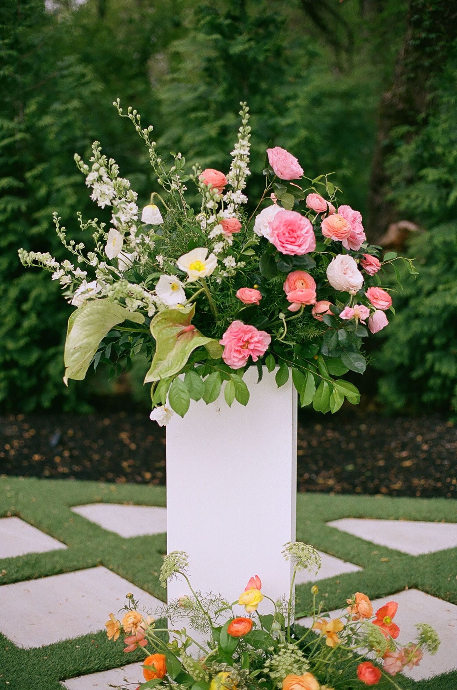 Elegant white pedestal floral arrangement with pink roses and white blooms in garden setting.
