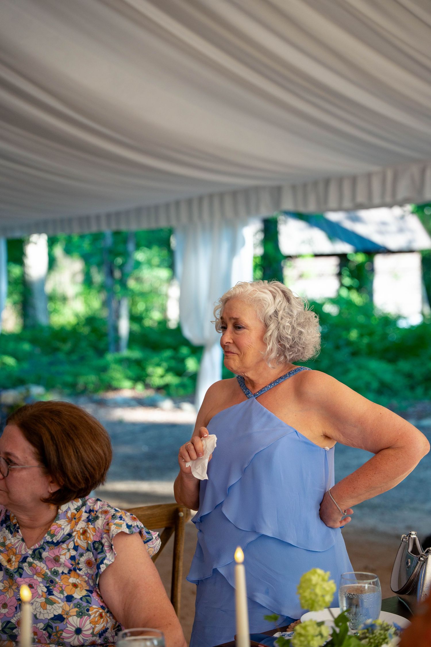 Elegant older woman in blue dress standing at outdoor wedding reception under white tent.