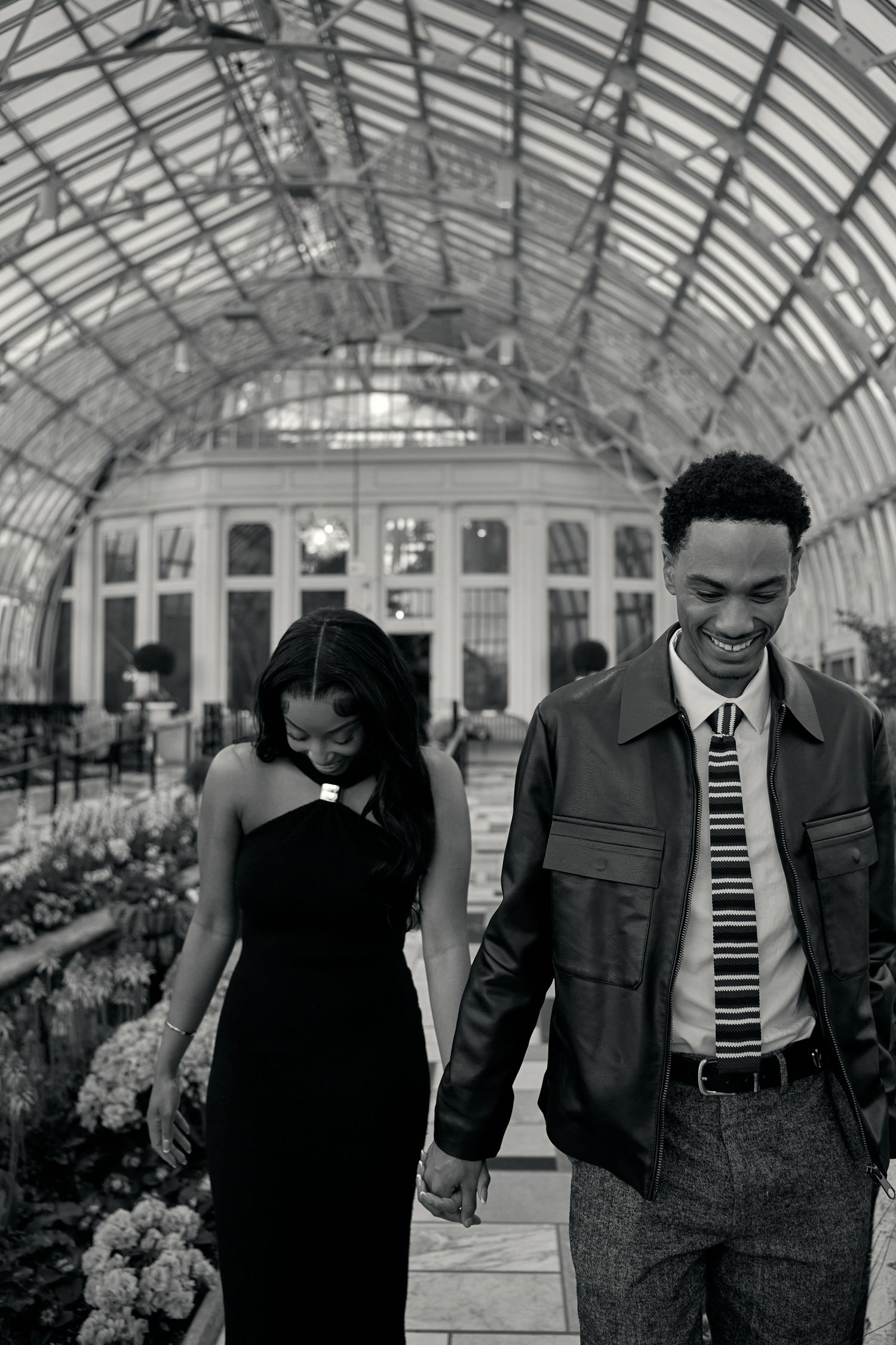 Black and white photo of engaged couple holding hands walking through an elegant glass greenhouse.