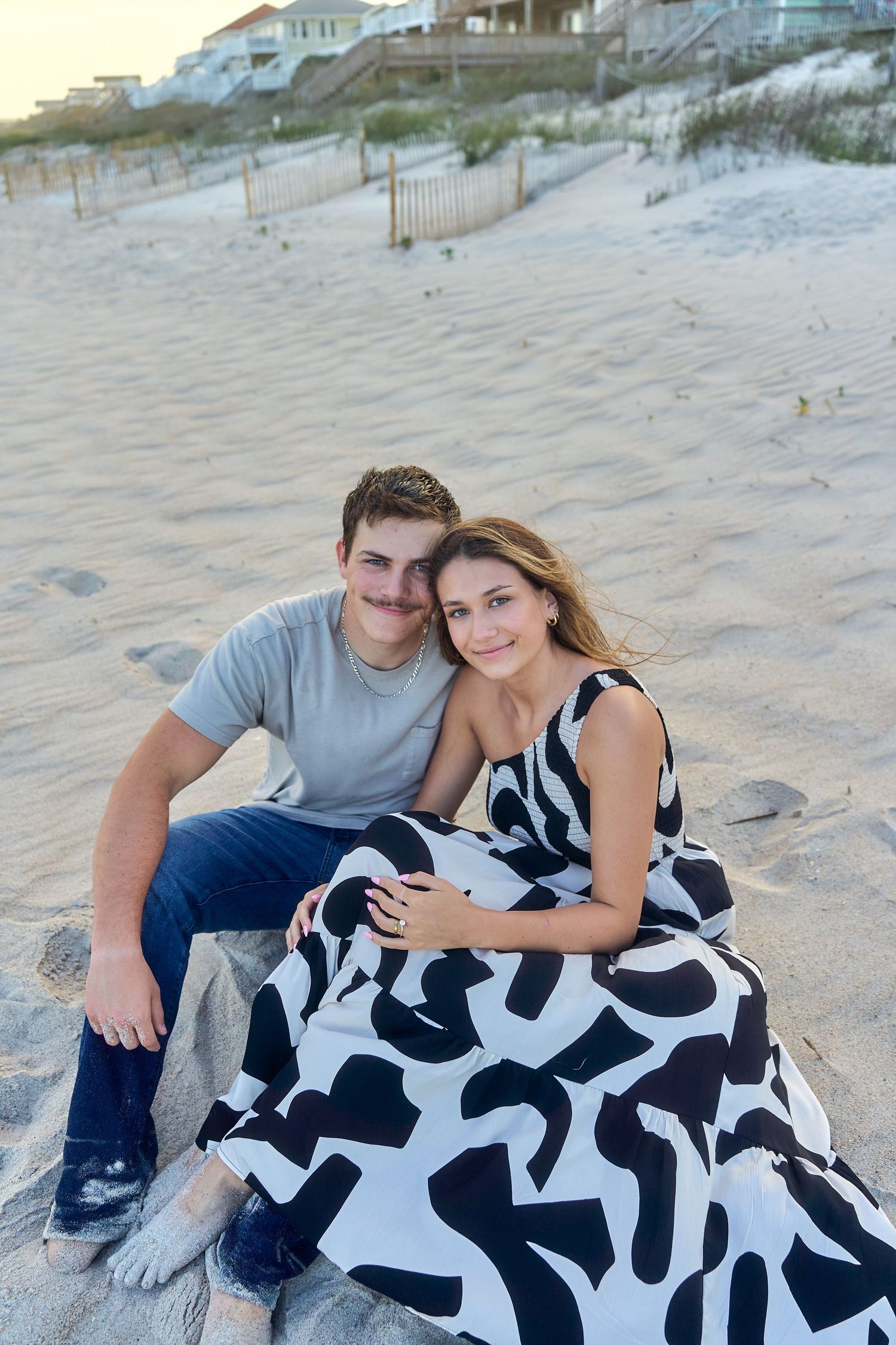 Couple sitting together on sandy beach at dusk with beach houses and dunes in background.