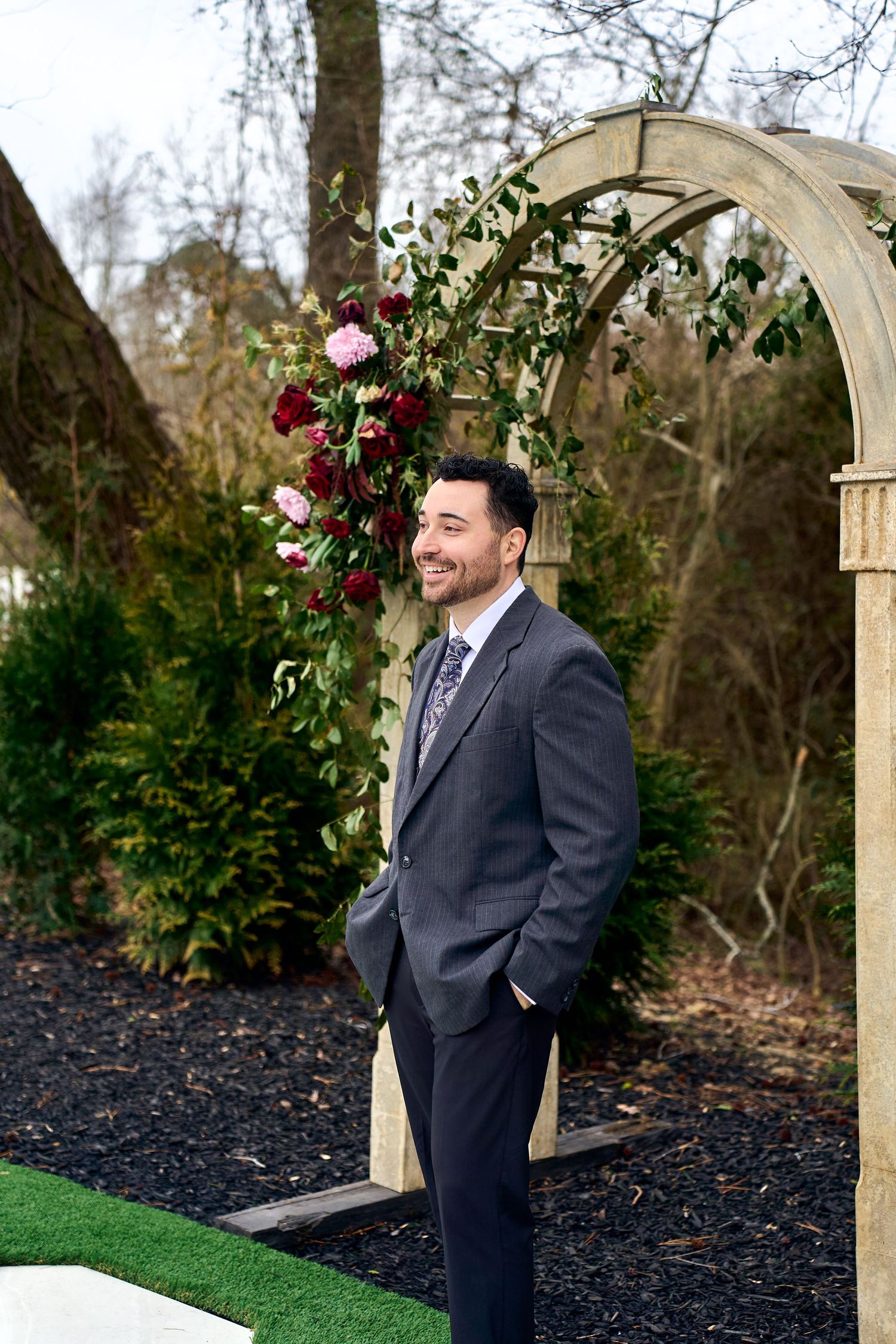 Well-dressed figure stands beside a garden archway decorated with pink and red climbing roses in an outdoor setting.