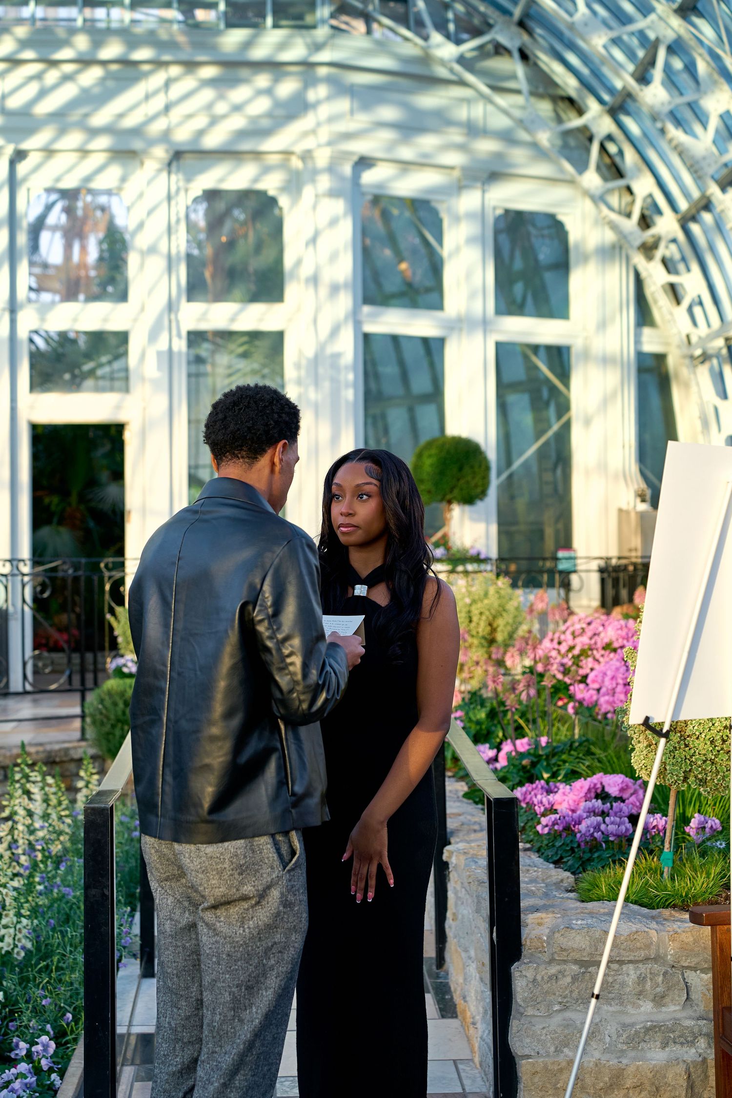 Couple in elegant black dress and leather jacket talking outside a glass greenhouse with colorful flowers.
