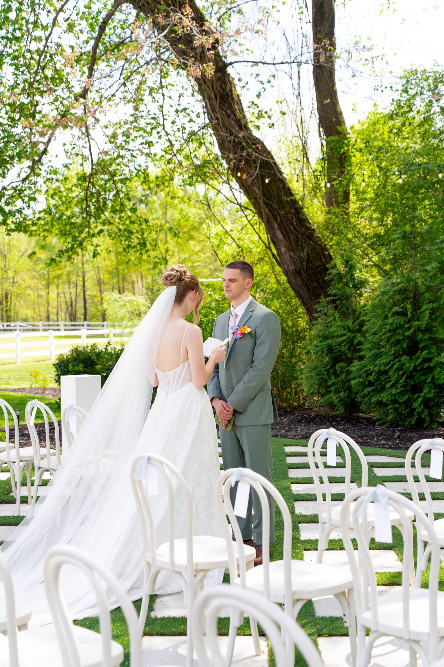 Bride and groom sharing a tender moment during outdoor wedding ceremony surrounded by white chairs and green trees.