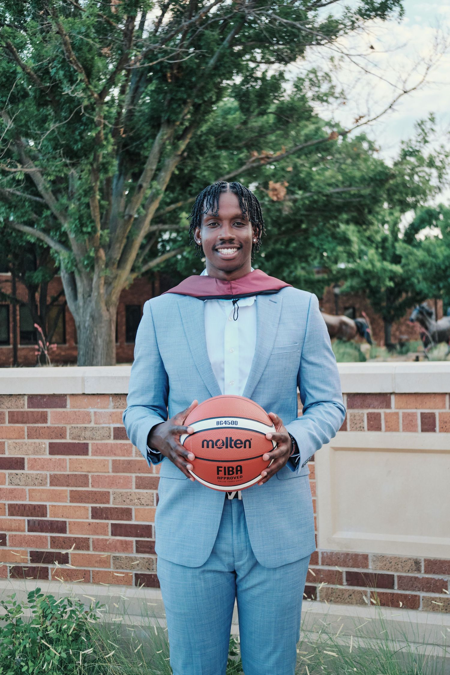 Smiling graduate in light blue suit holding a Molten FIBA basketball outdoors on campus.