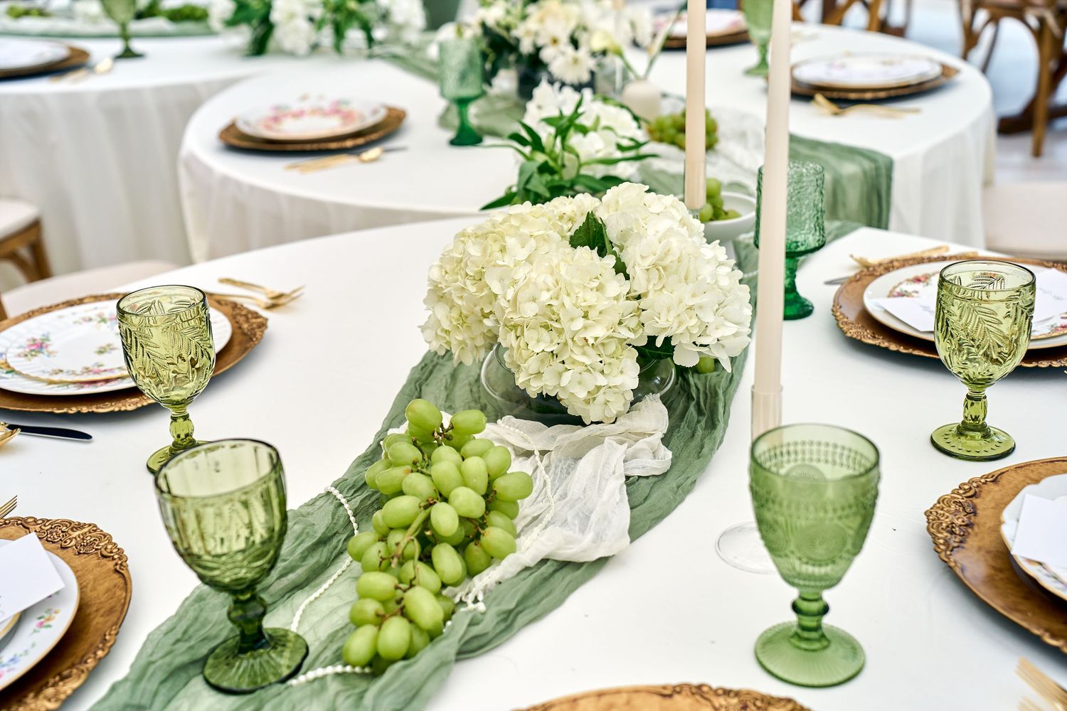 Elegant table setting with green glassware, white hydrangeas, fresh grapes and sage green table runner atop white tablecloth.
