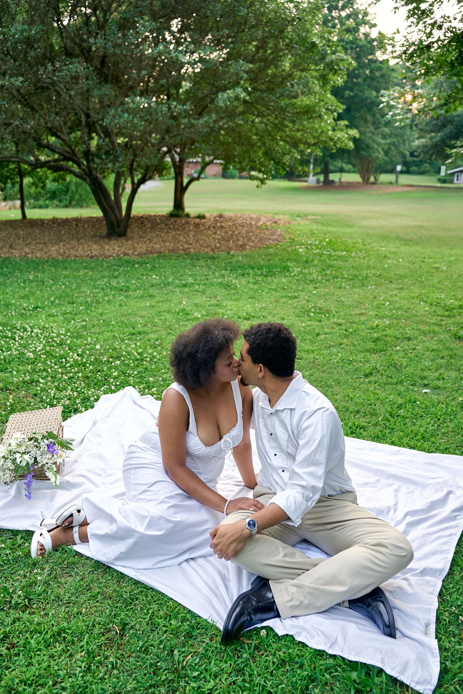 Pair shares intimate moment while relaxing on white blanket in peaceful park setting surrounded by trees.