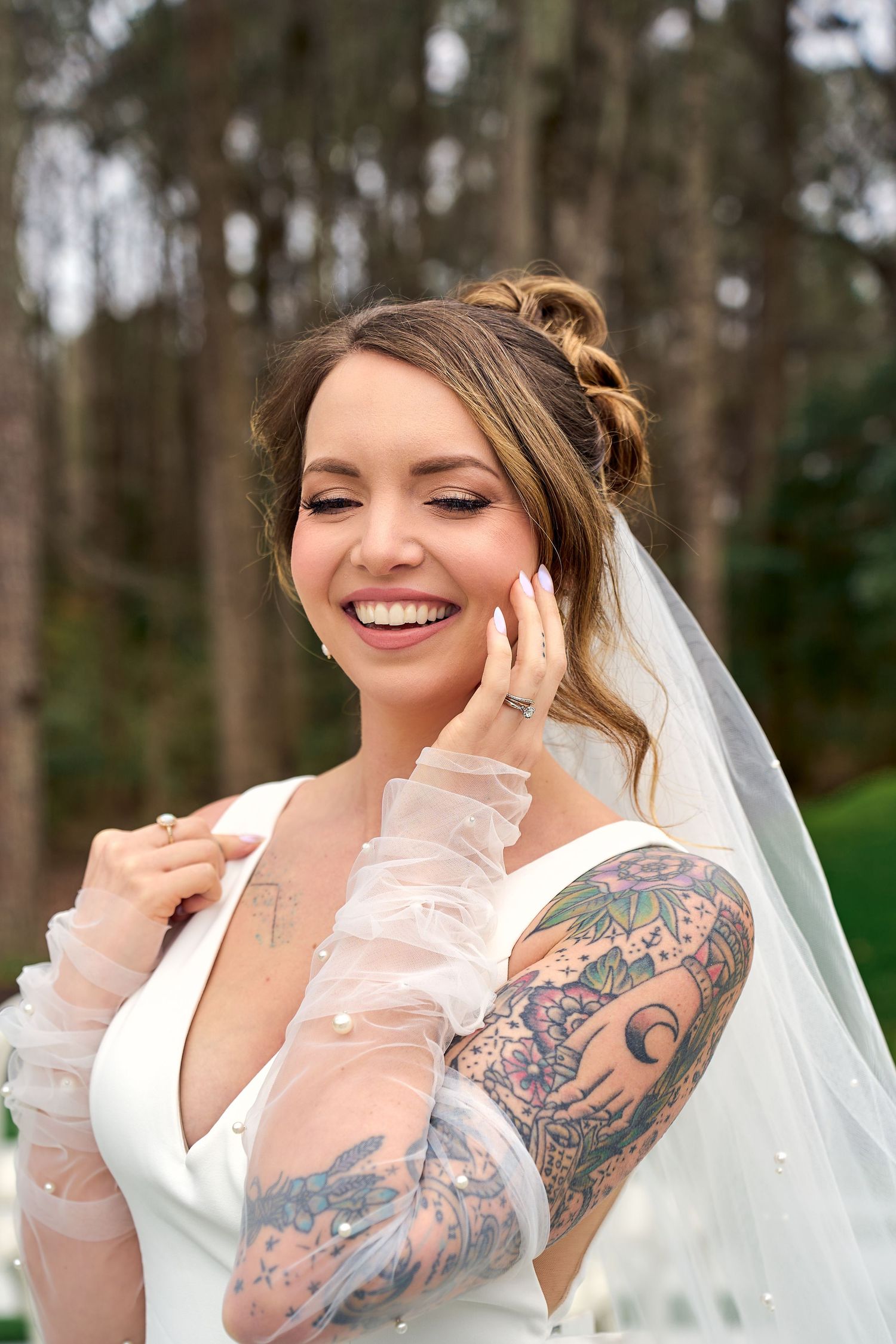 A bride in a white wedding dress with visible tattoo sleeves poses outdoors in a forest setting while wearing a veil.