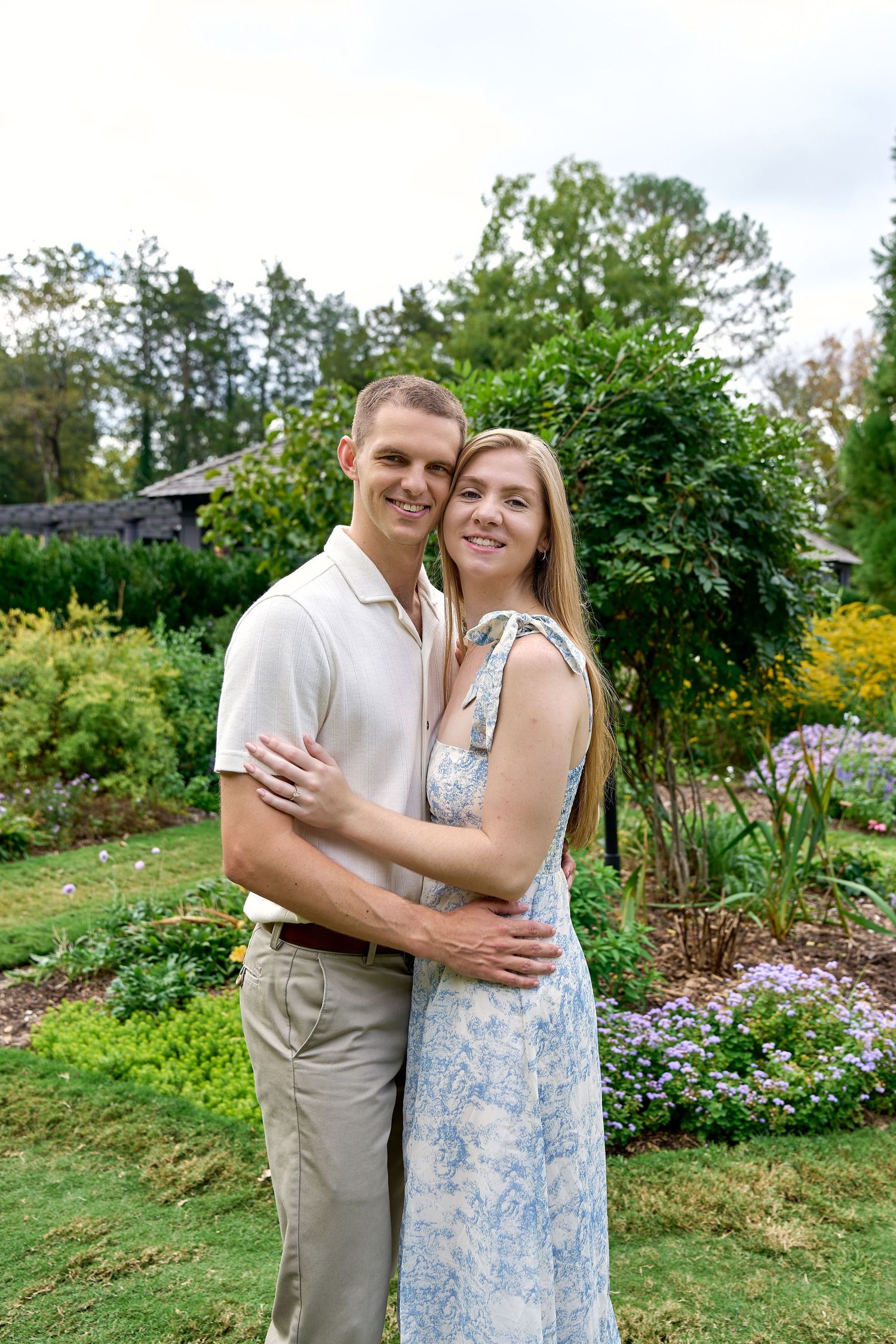 Smiling couple embracing in a lush garden with colorful flowers and green trees.