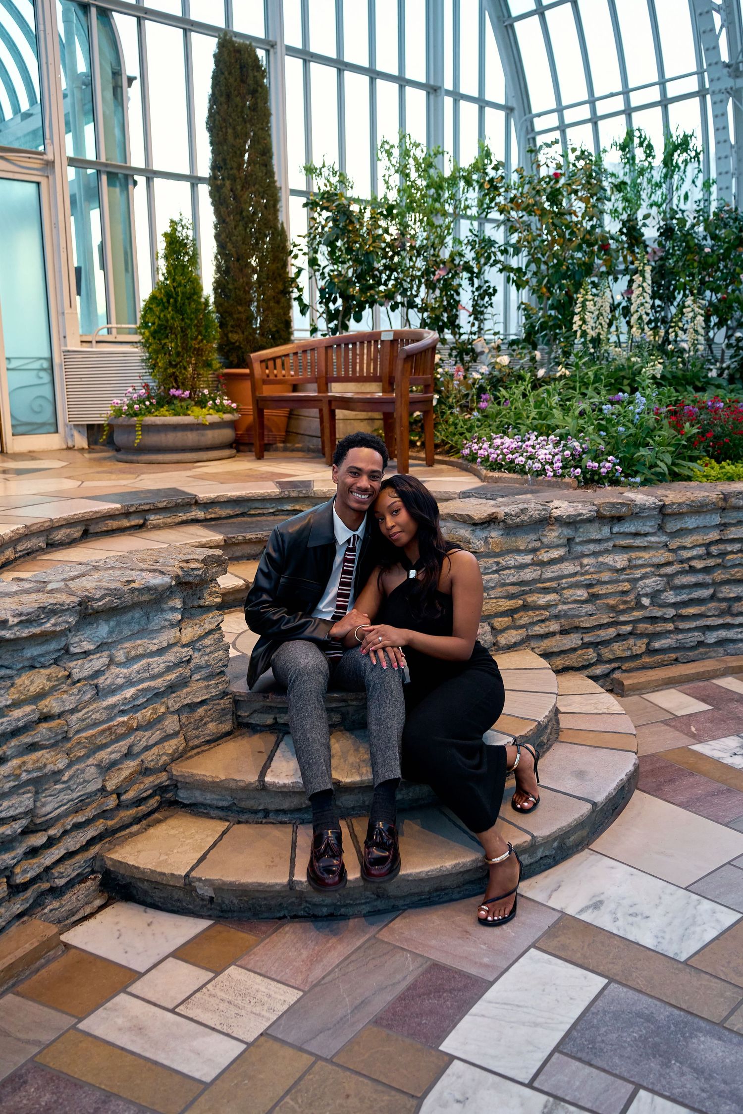 Engaged couple sitting together on stone steps inside a lush indoor botanical garden conservatory.