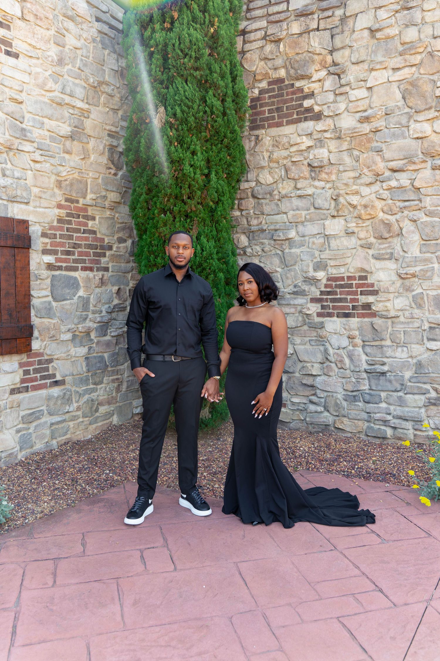 Stylish couple in coordinating black outfits posing together against rustic stone wall with climbing evergreen plant.