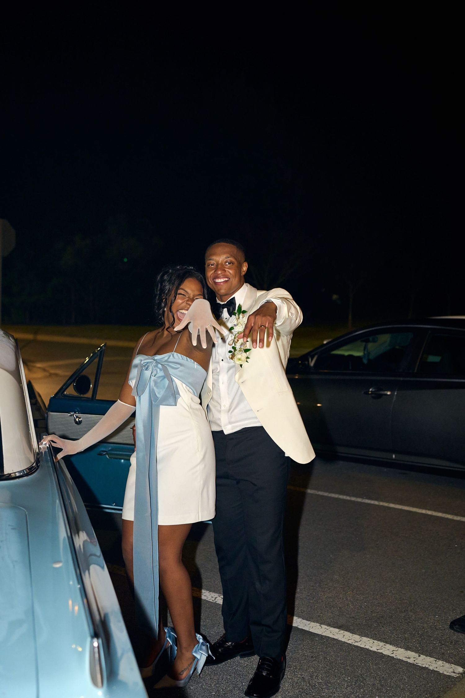 Bride and groom embrace and laugh beside a vintage light blue classic car after their wedding.