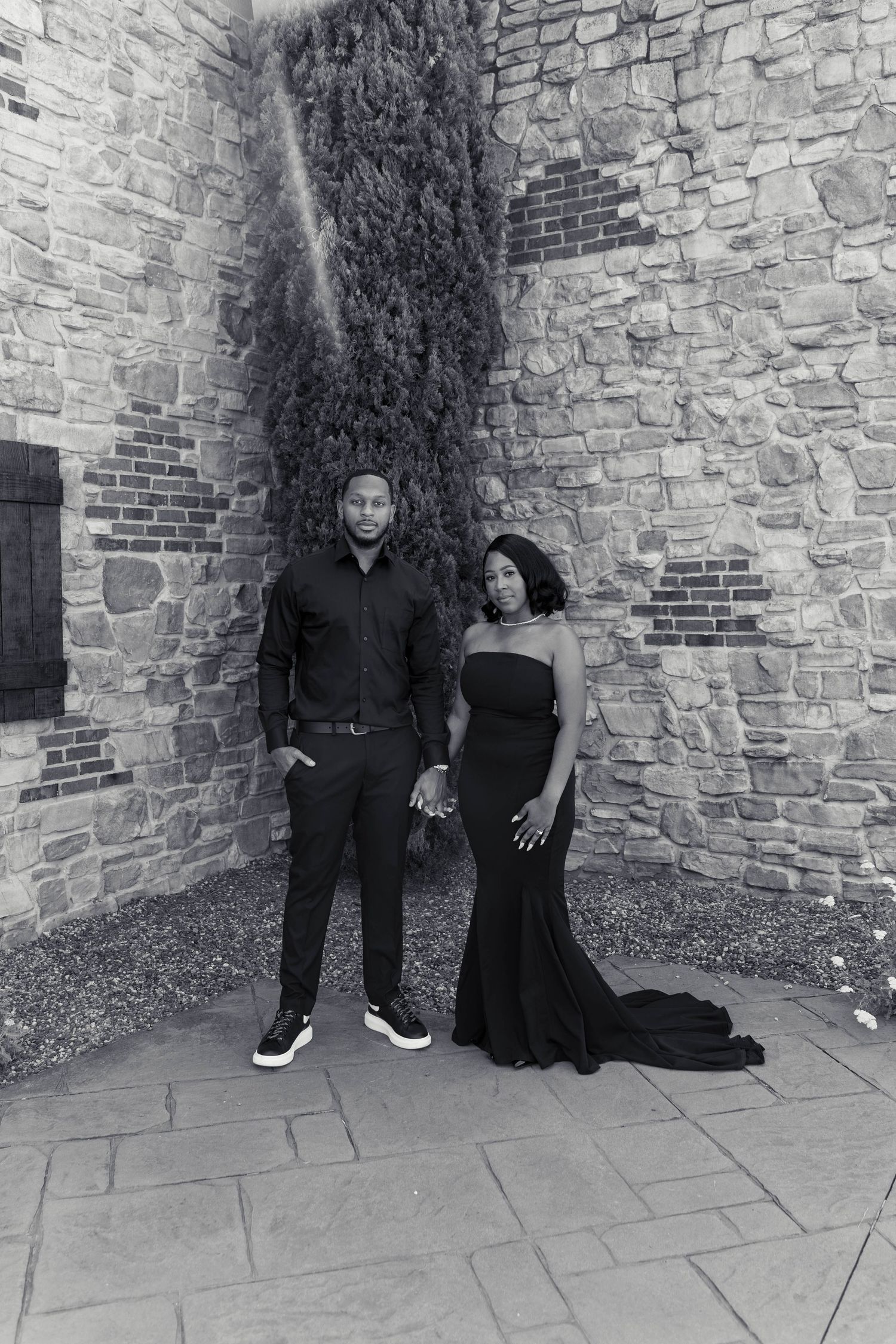 Elegant couple in matching black outfits posing against a rustic stone wall in a black and white portrait.