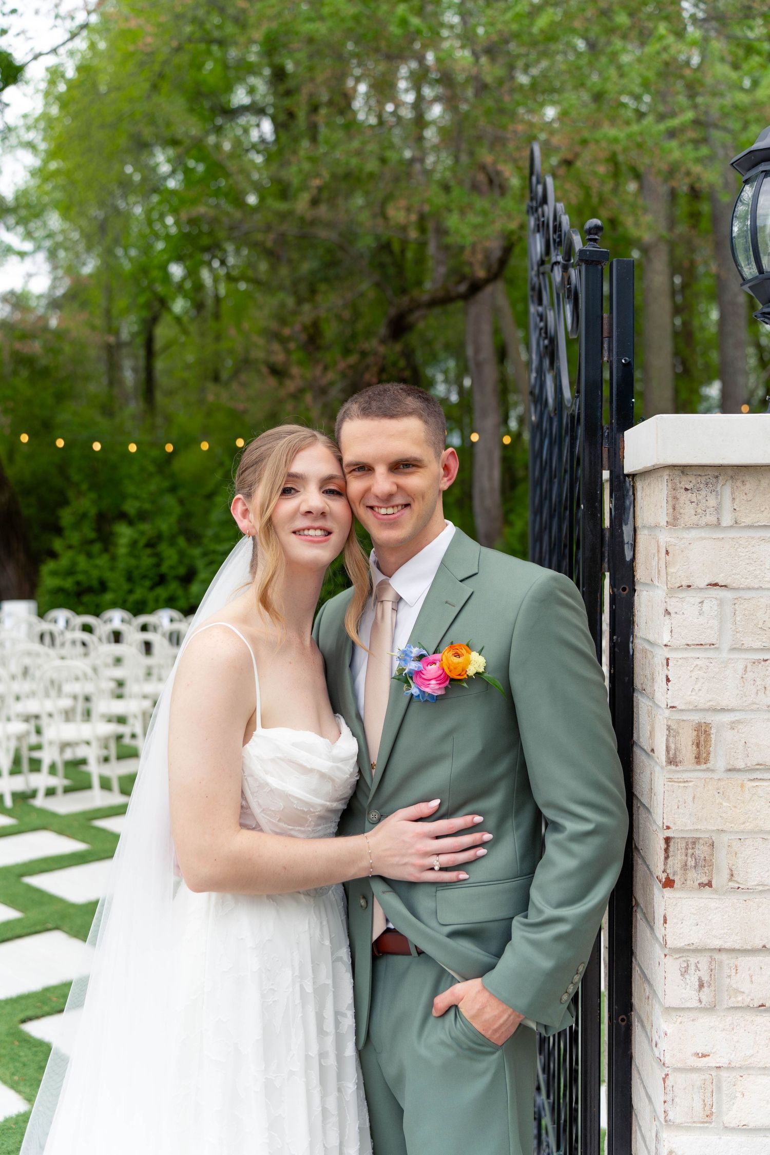 Bride in white gown and groom in sage green suit pose together at an outdoor wedding venue gate.