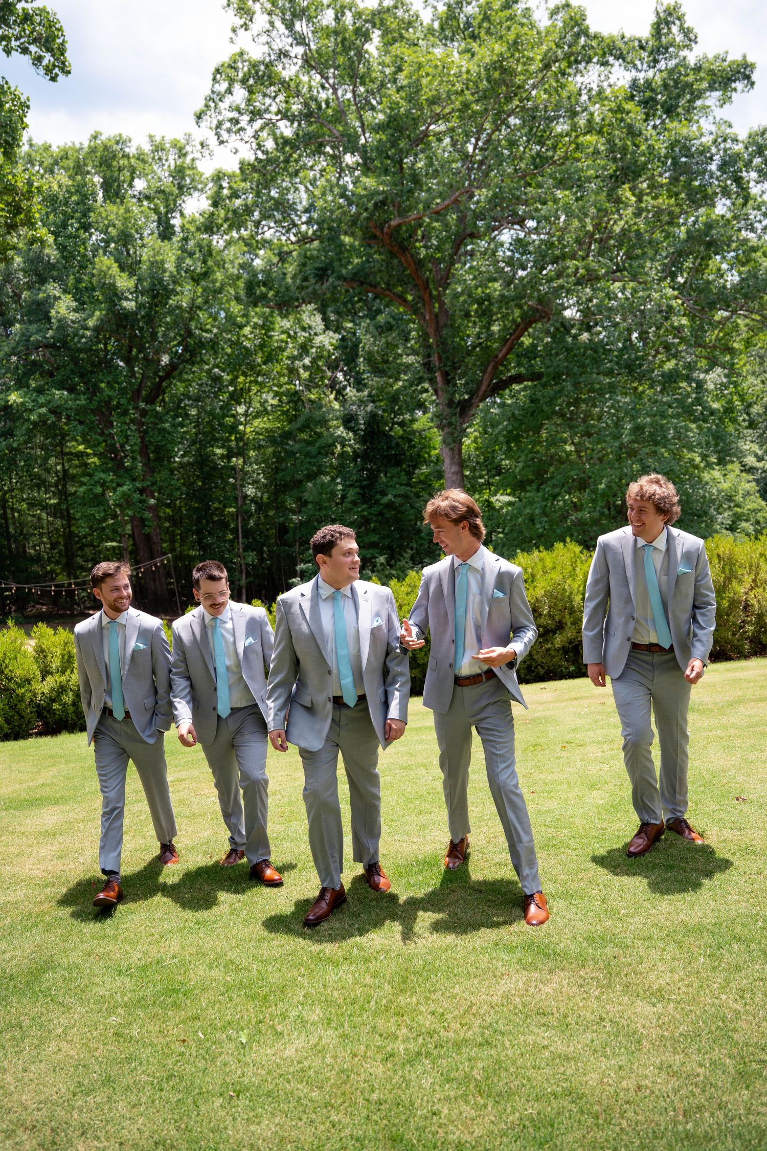 Five groomsmen in matching gray suits with mint ties walk across a green lawn at a wedding.