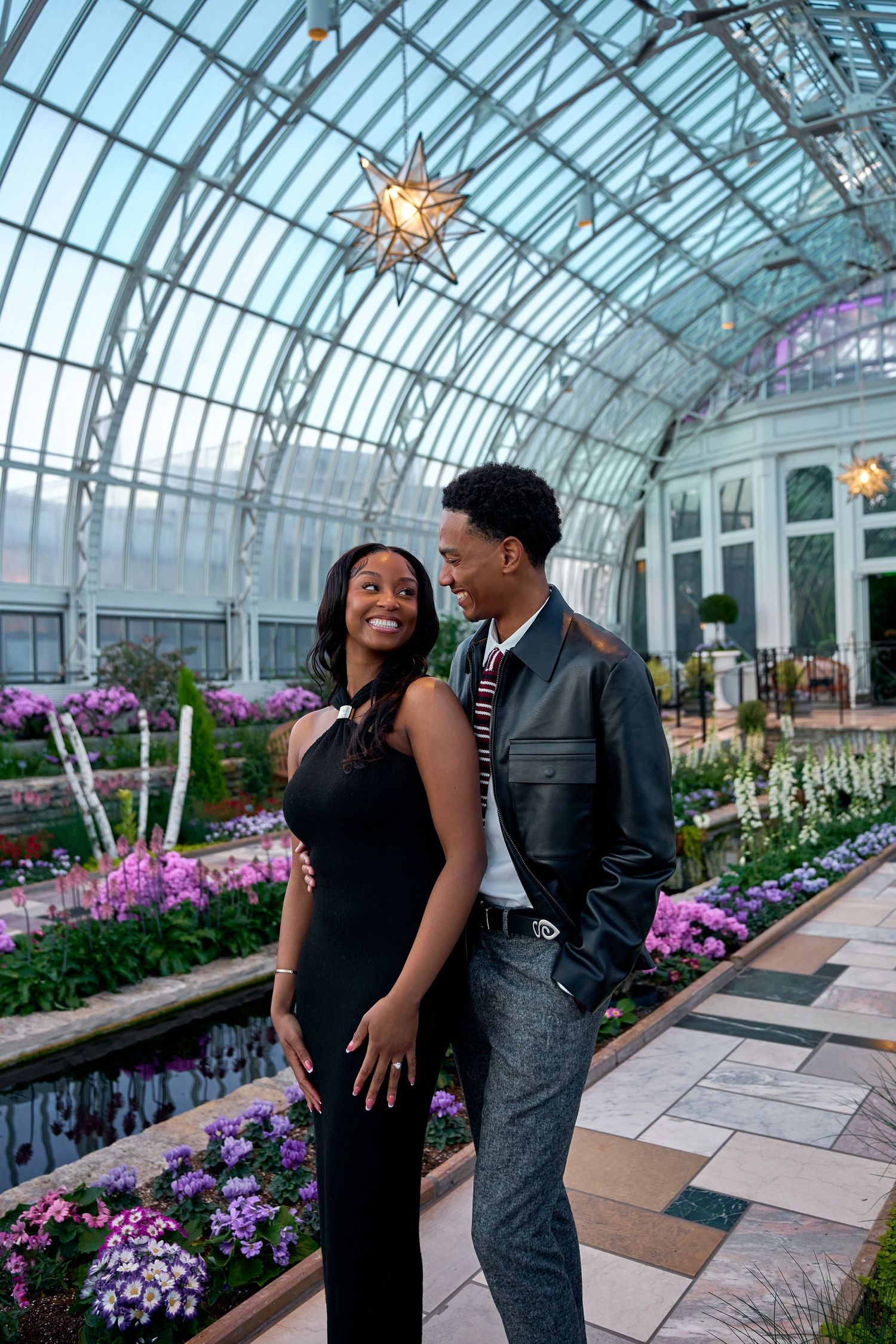 Happy engaged couple in stylish outfits smiling at each other inside a stunning botanical conservatory.