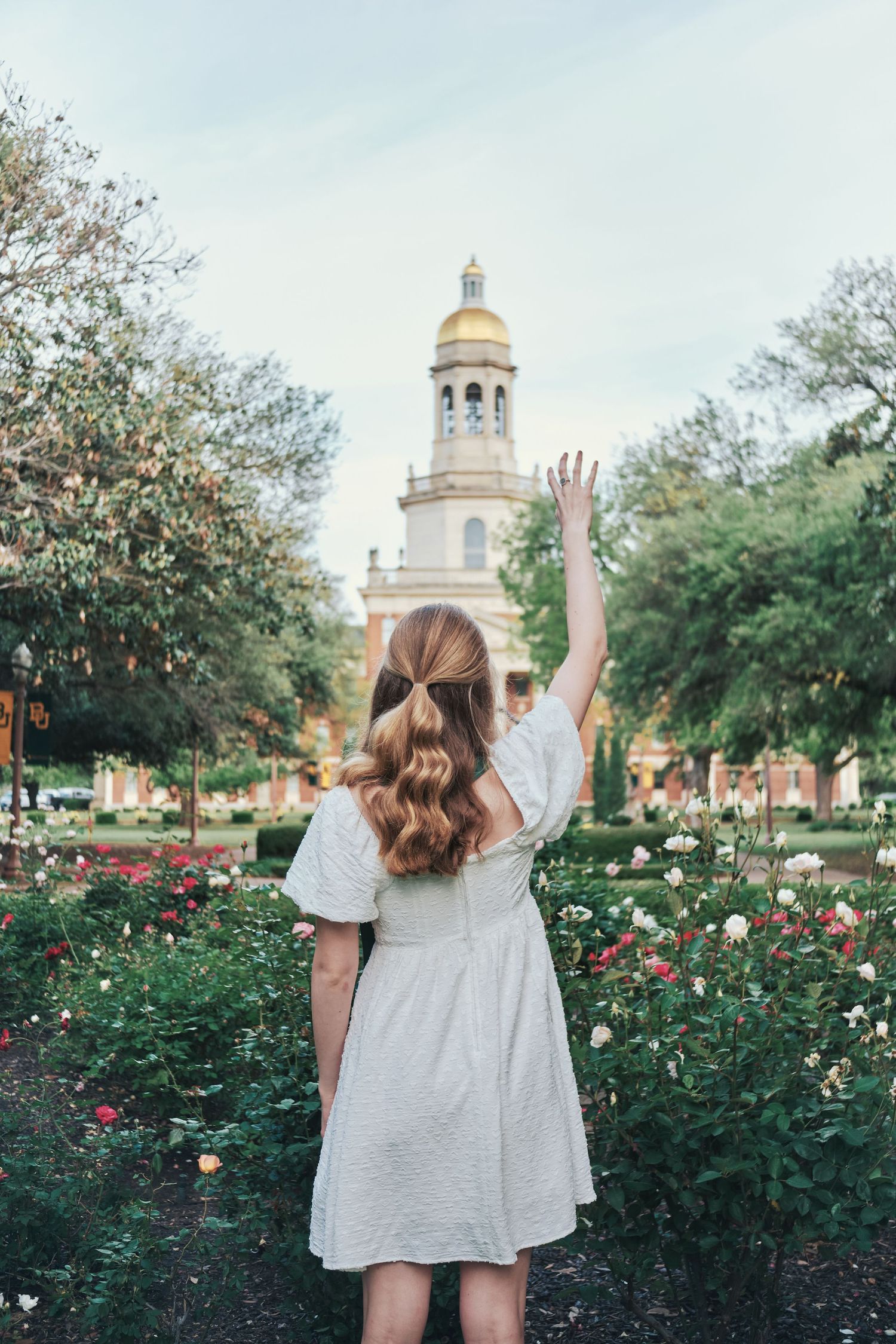 Woman in white dress waving toward Baylor University chapel surrounded by rose garden. North Carolina Photographer.