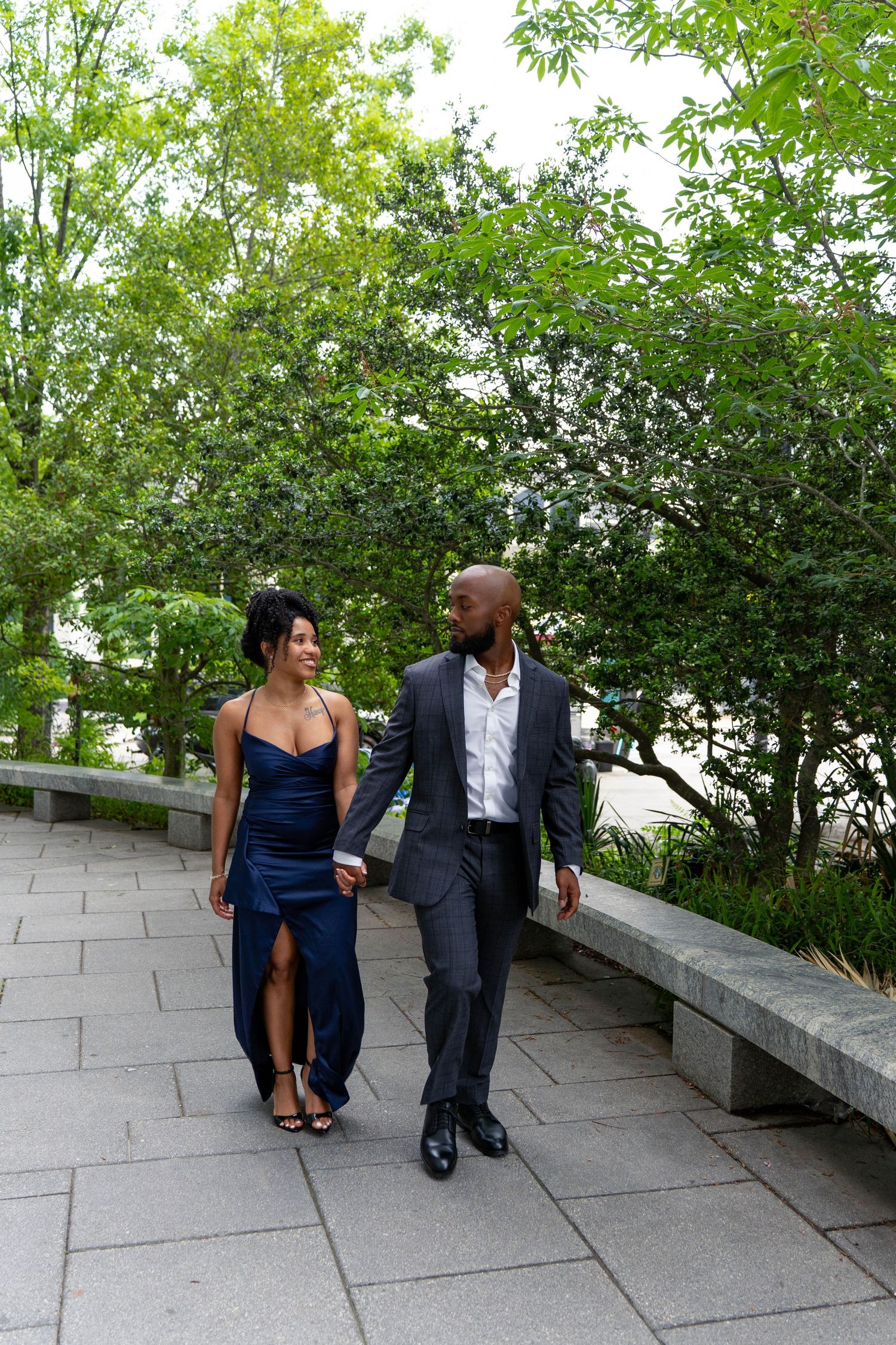 Happy Black couple holding hands strolling along a tree-lined path, dressed in elegant formal attire.