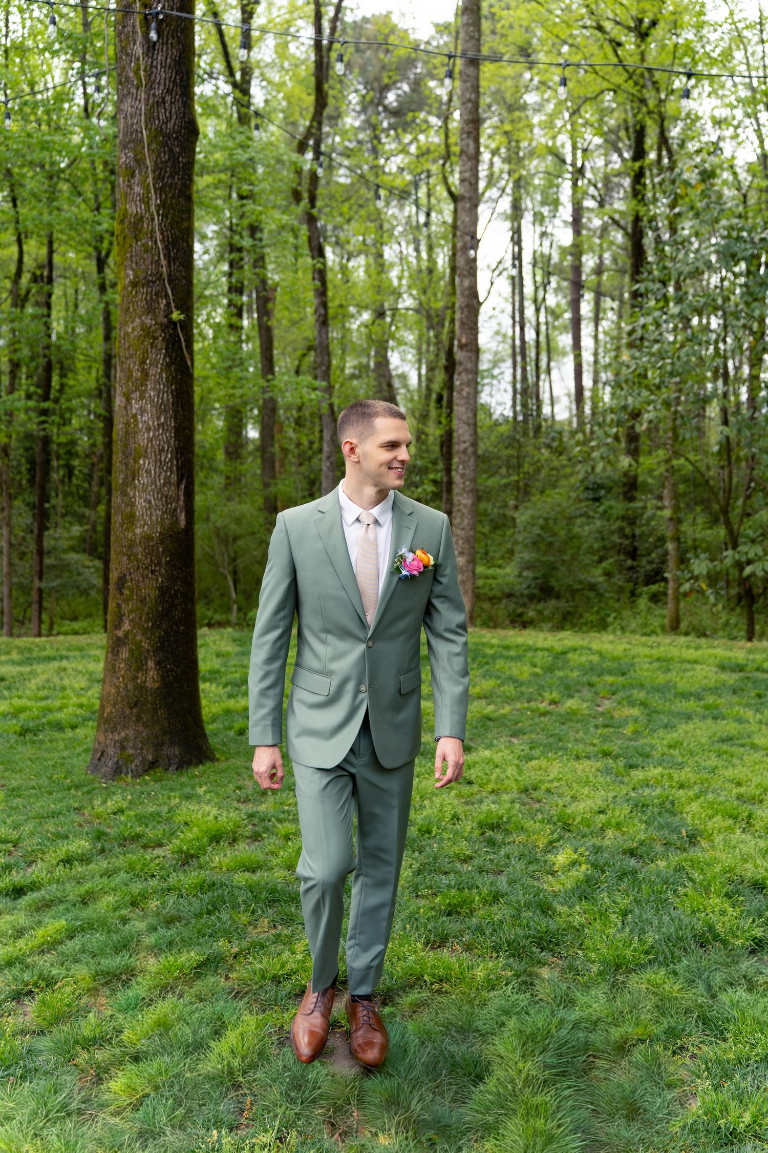 Groom in sage green suit with colorful boutonniere standing in lush green forest setting.