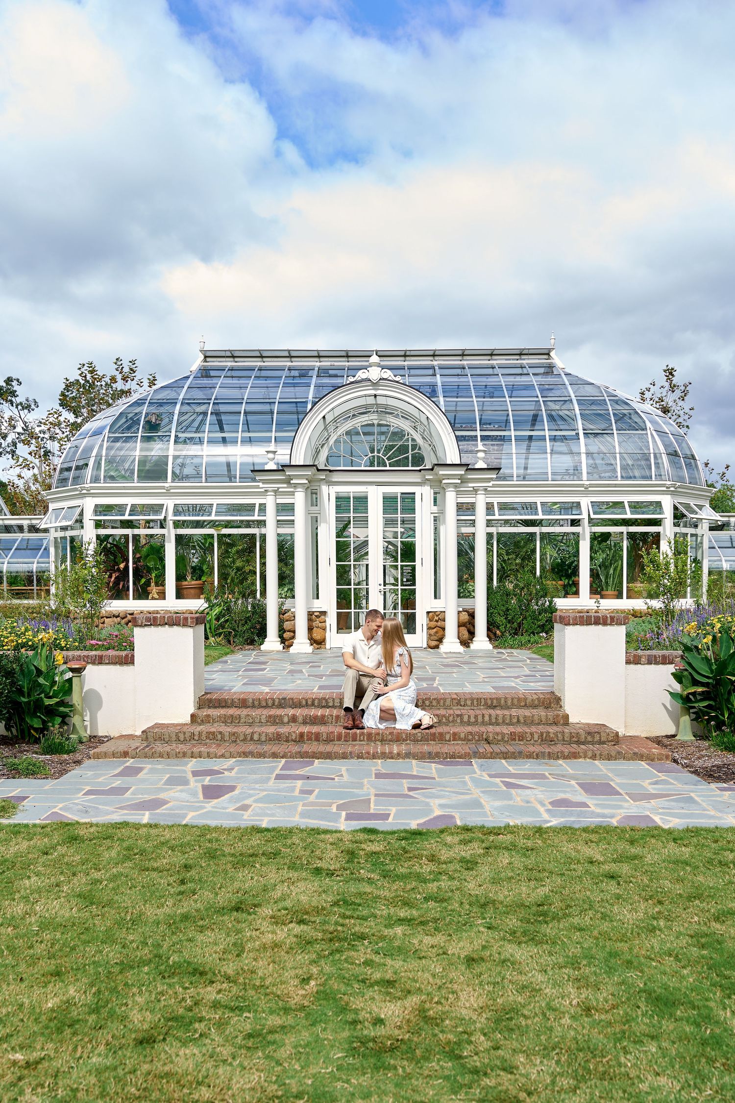 Couple sitting on steps in front of a beautiful white glass greenhouse conservatory with manicured gardens.