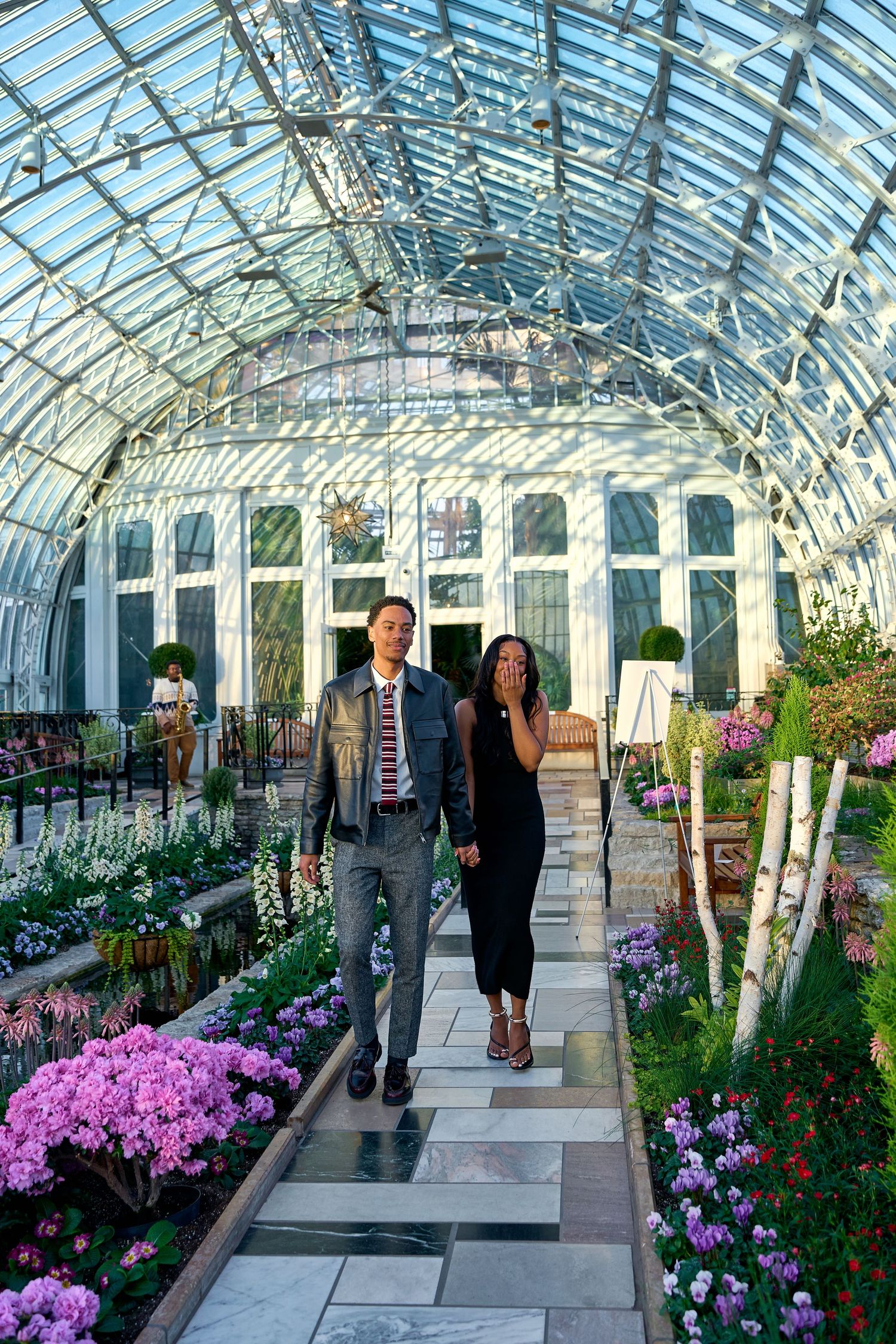 Engaged couple walking hand in hand through a stunning glass conservatory filled with vibrant pink flowers.
