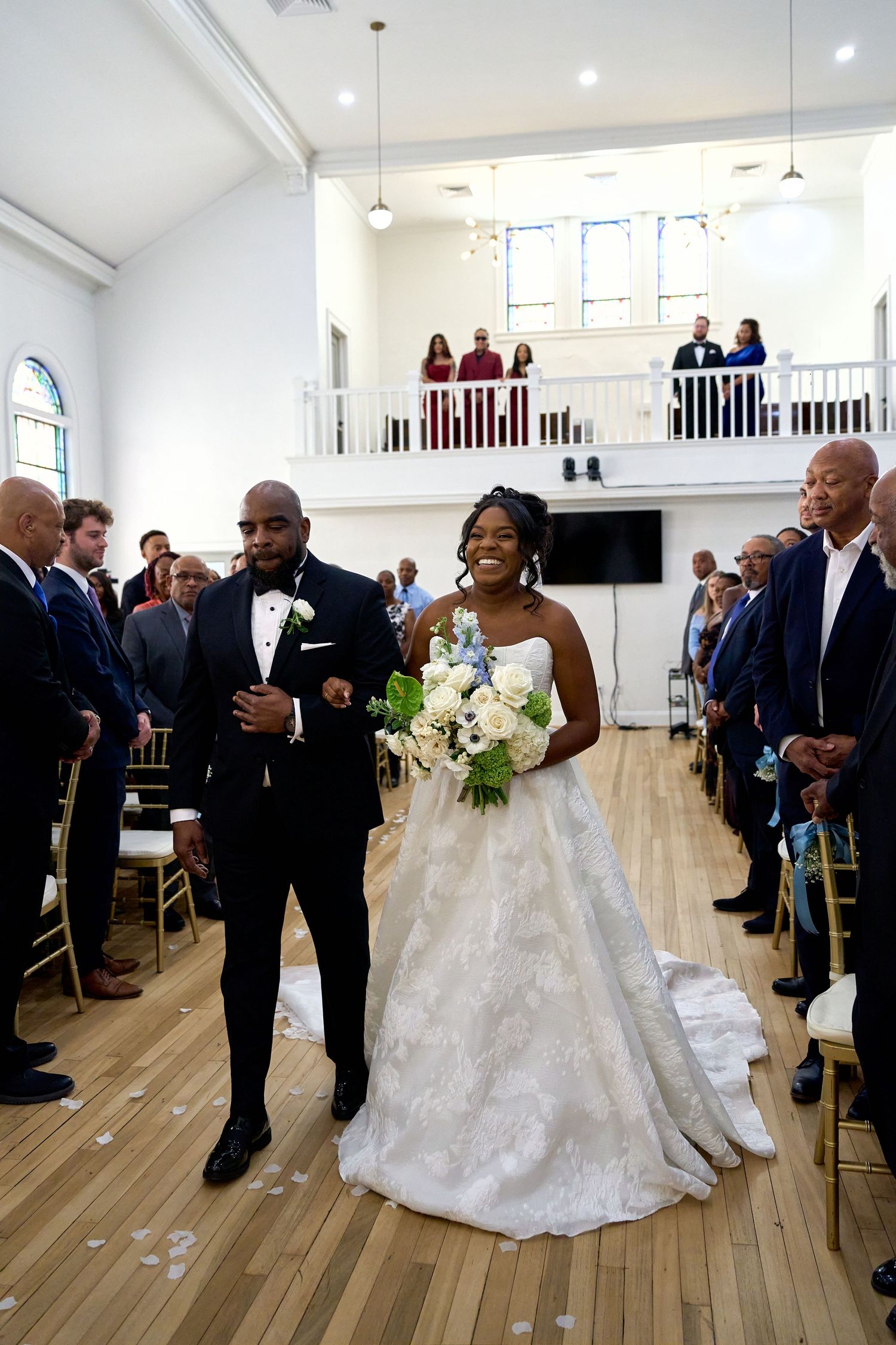 Smiling bride walks down aisle in lace ballgown holding white bouquet during church wedding ceremony.