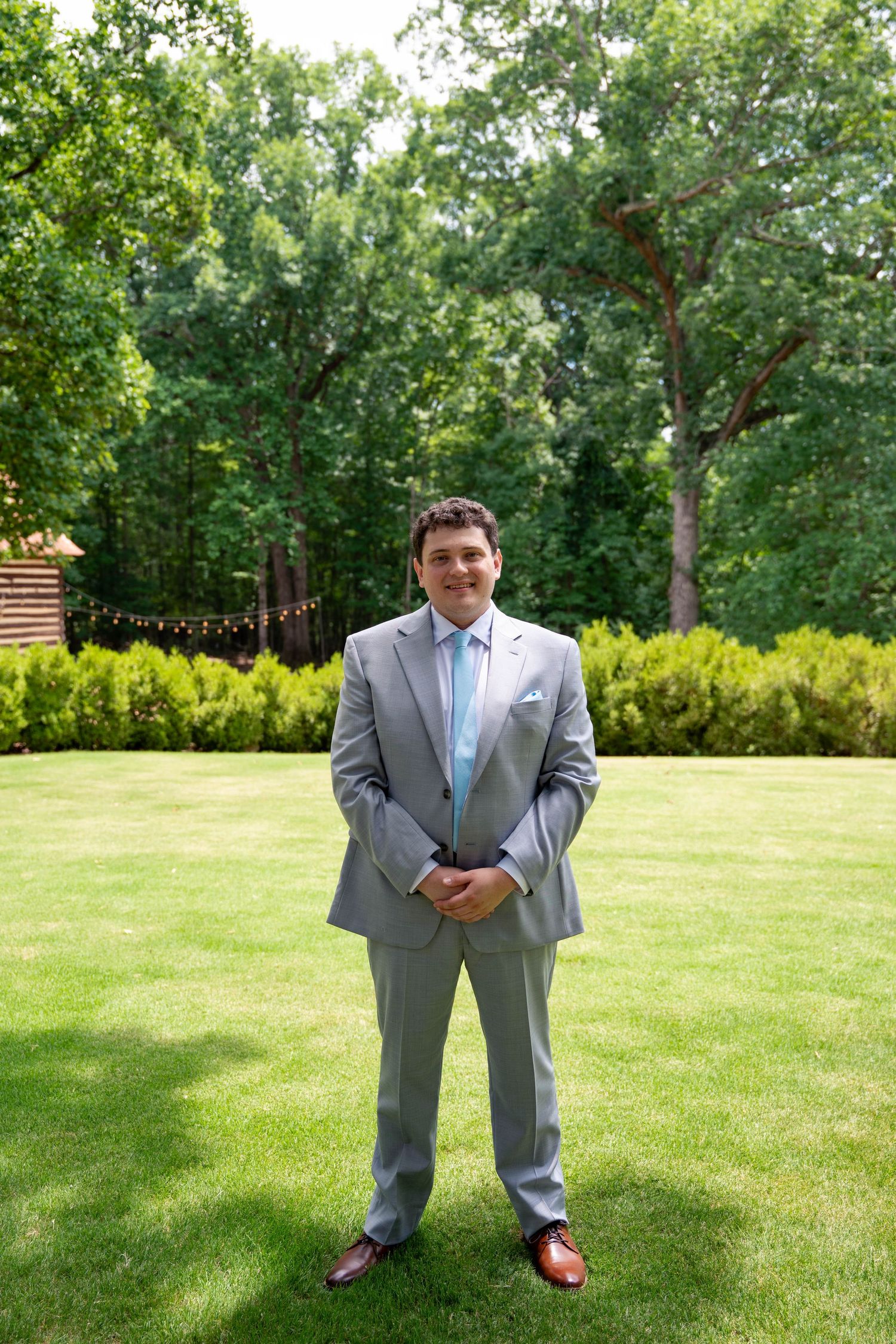 Groom in gray suit and blue tie standing on green lawn at outdoor wedding venue.