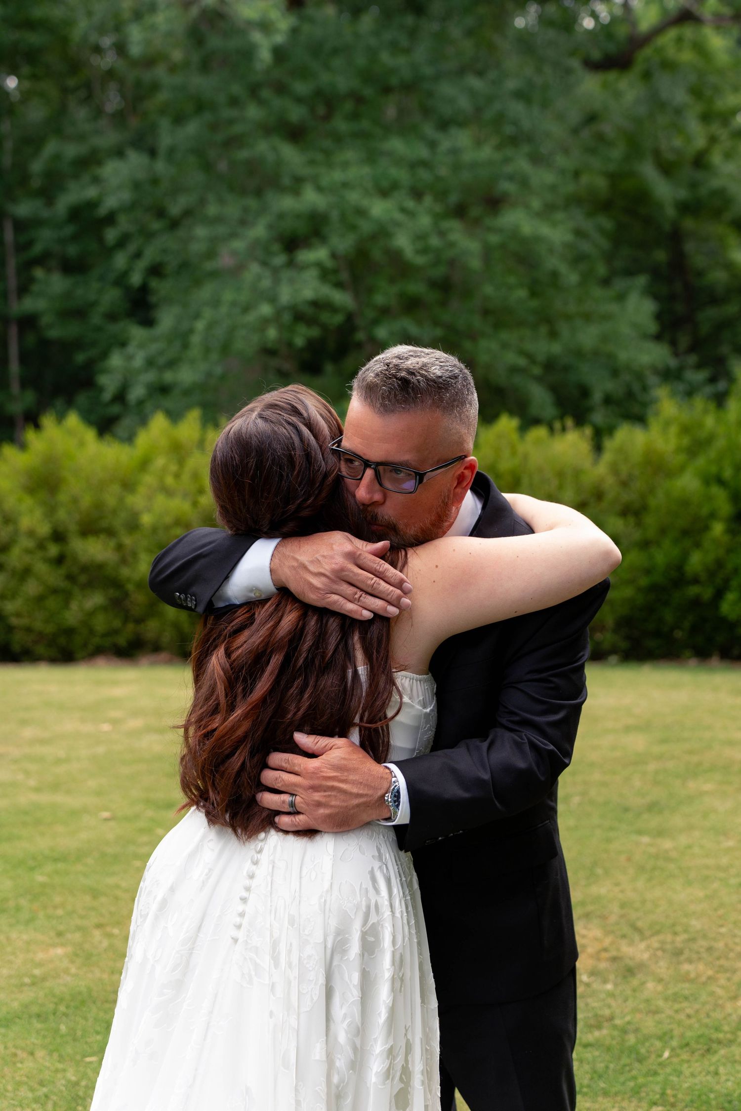 Emotional bride and older gentleman share a heartfelt embrace on the green lawn at an outdoor wedding.