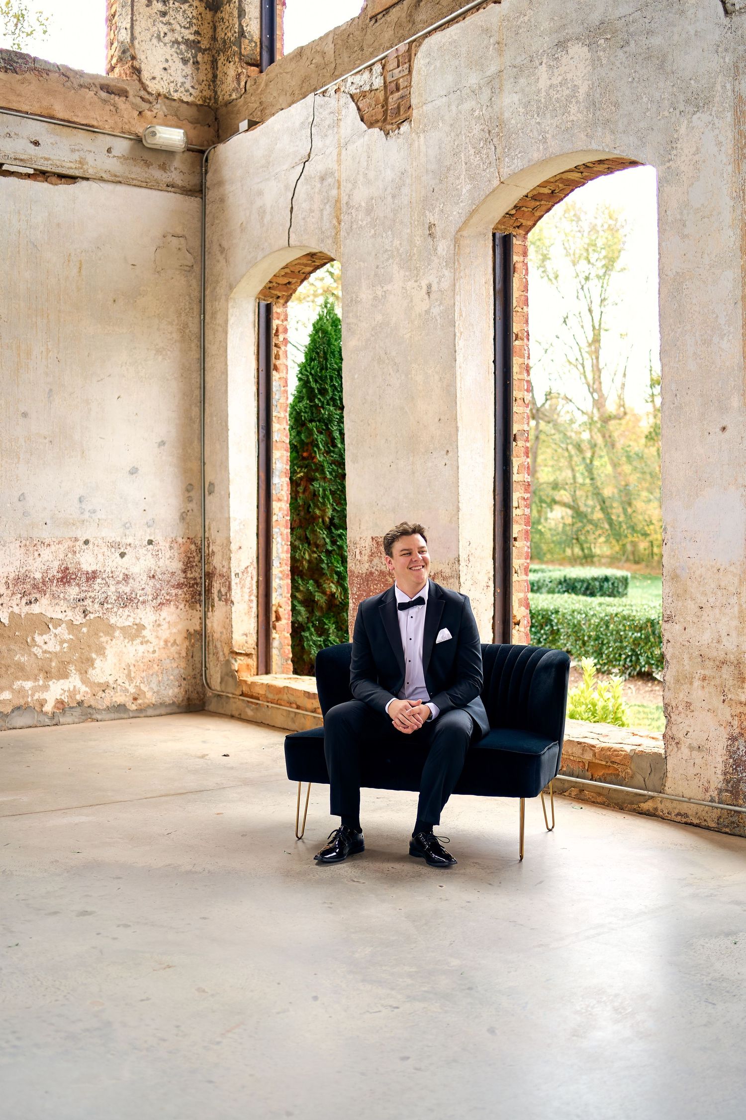 Person in formal black suit sits on modern armchair inside rustic stone building with arched windows overlooking garden.