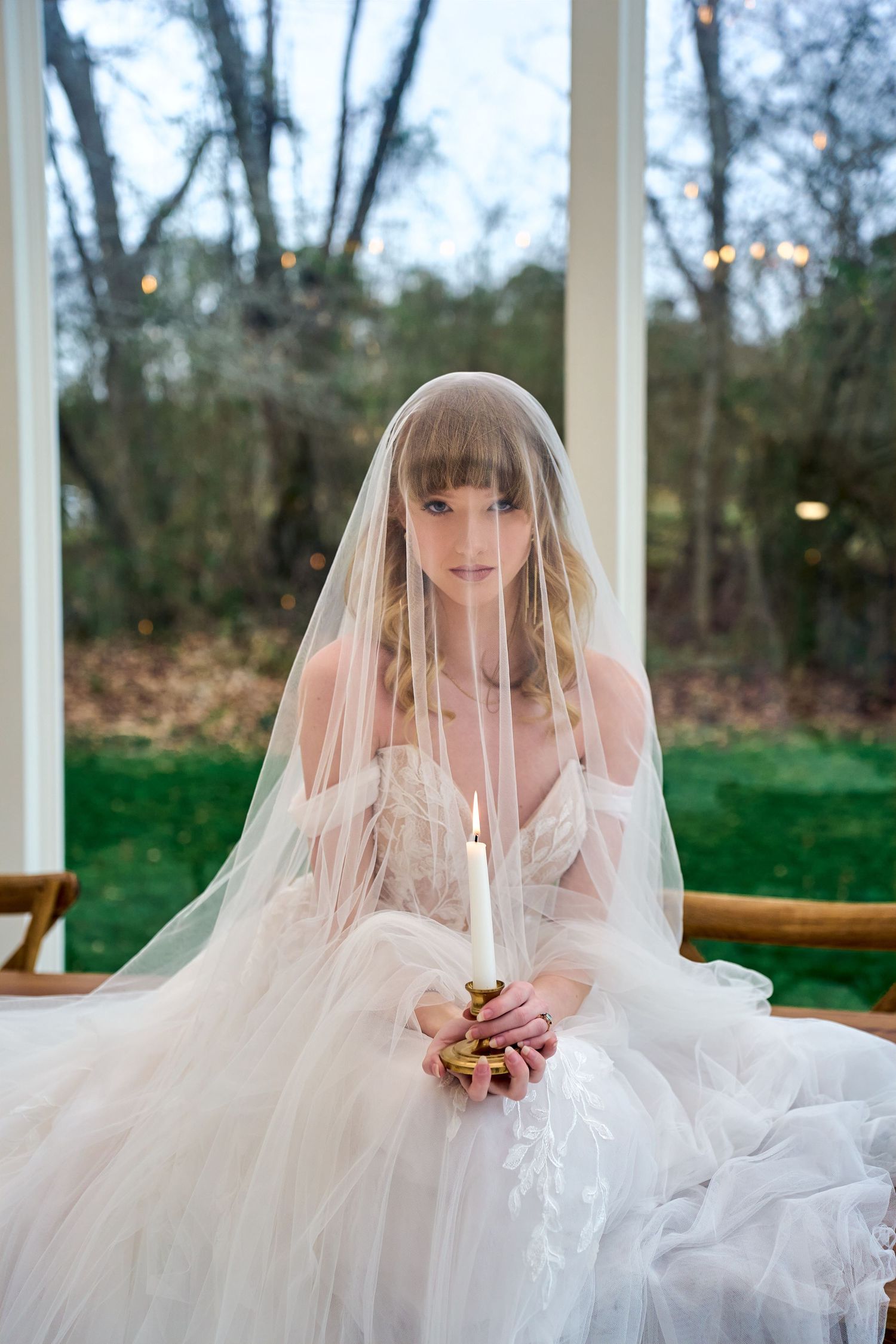 Ethereal bride in flowing white tulle gown and veil sits gracefully with clasped hands against woodland backdrop.