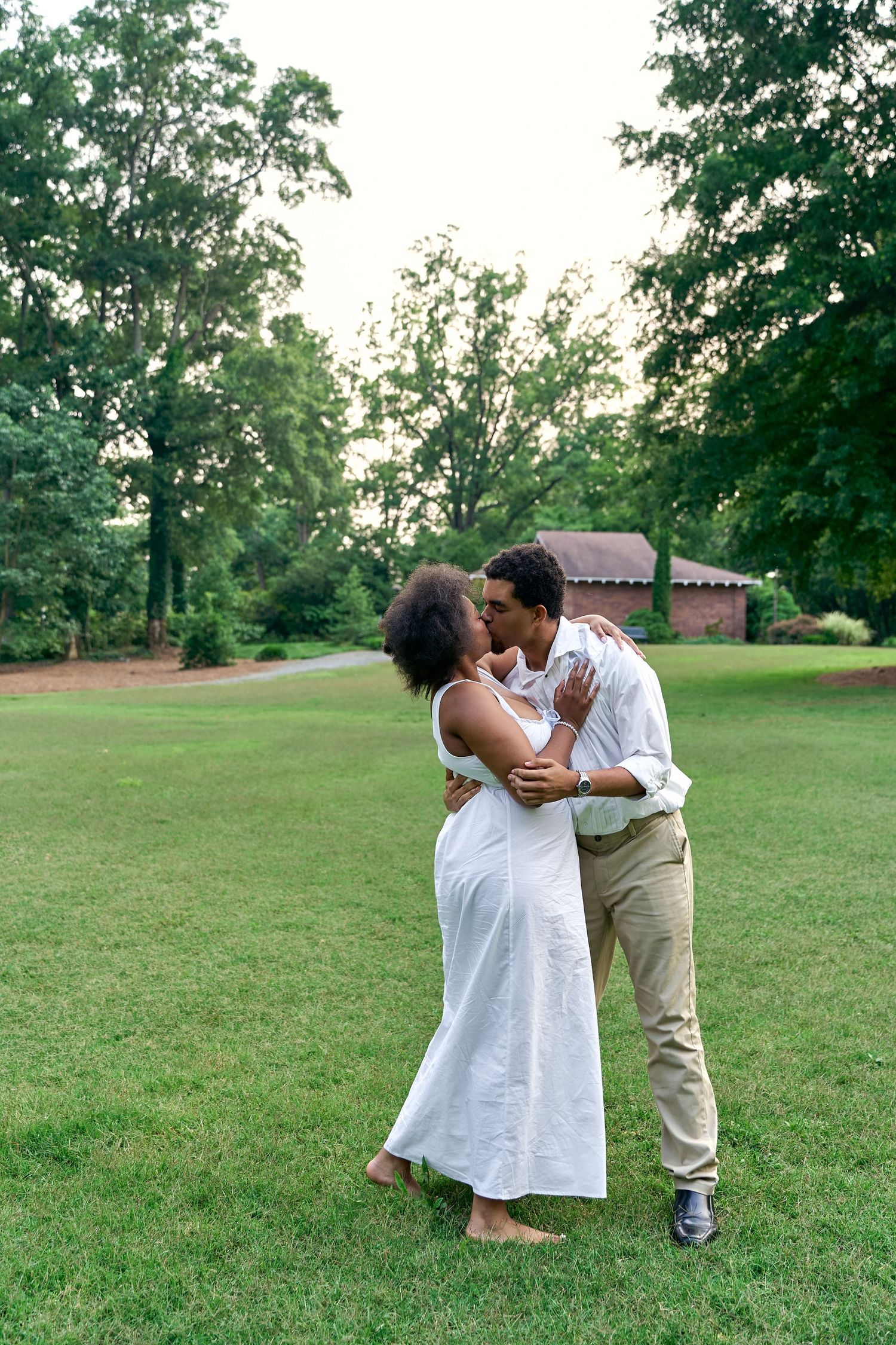 A couple embraces and kisses in a serene outdoor park setting with green grass and trees in the background.