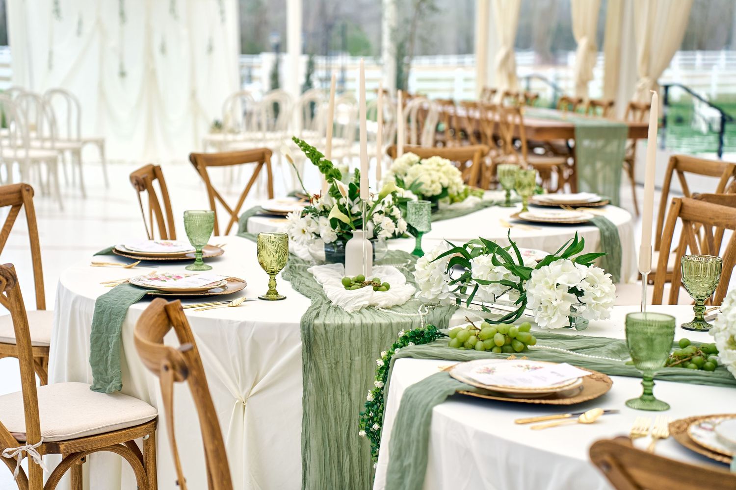An elegant wedding table setting with sage green runners, white linens and wooden cross-back chairs in a bright venue.