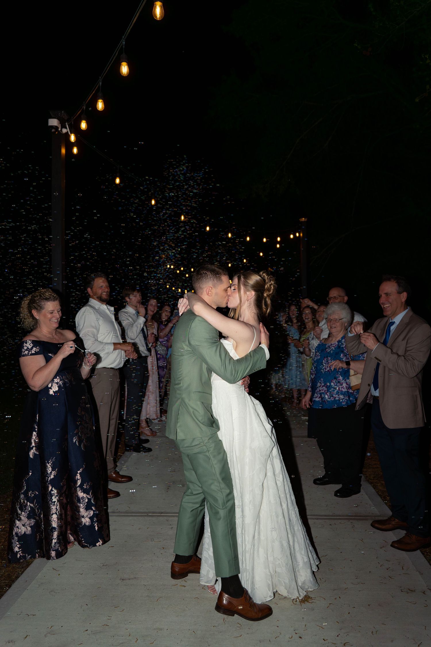 Newlyweds share a romantic kiss surrounded by cheering wedding guests under string lights at night.