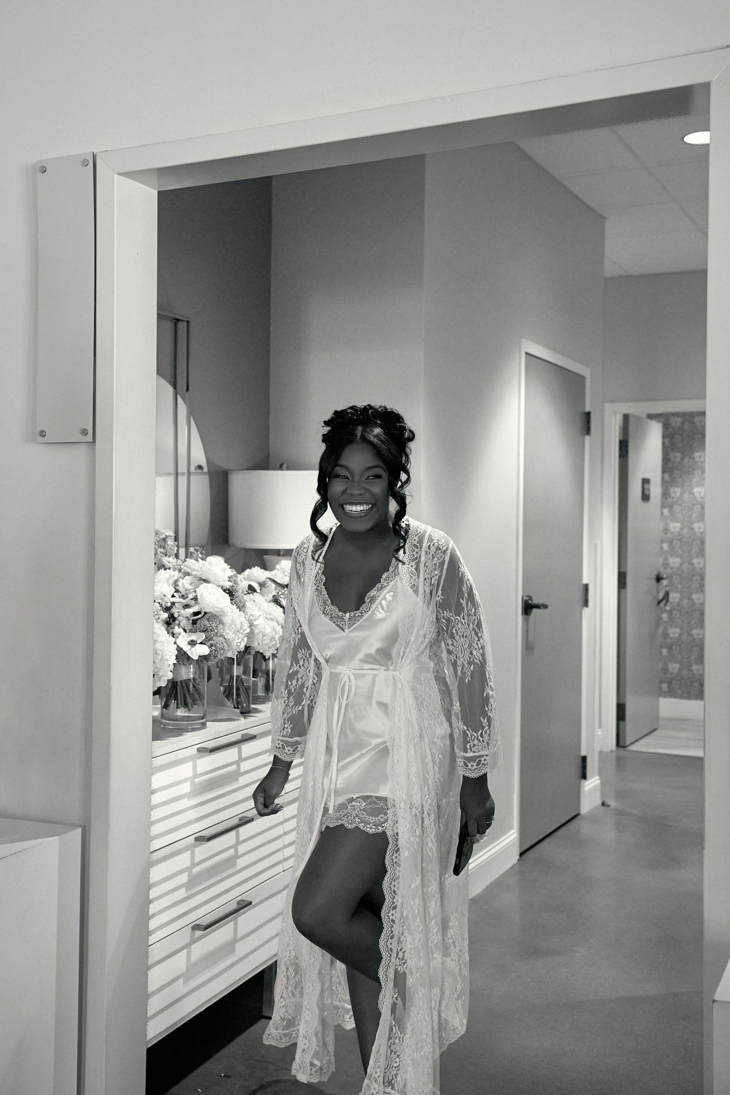 Smiling bride in lace robe getting ready in bridal suite in black and white photo.