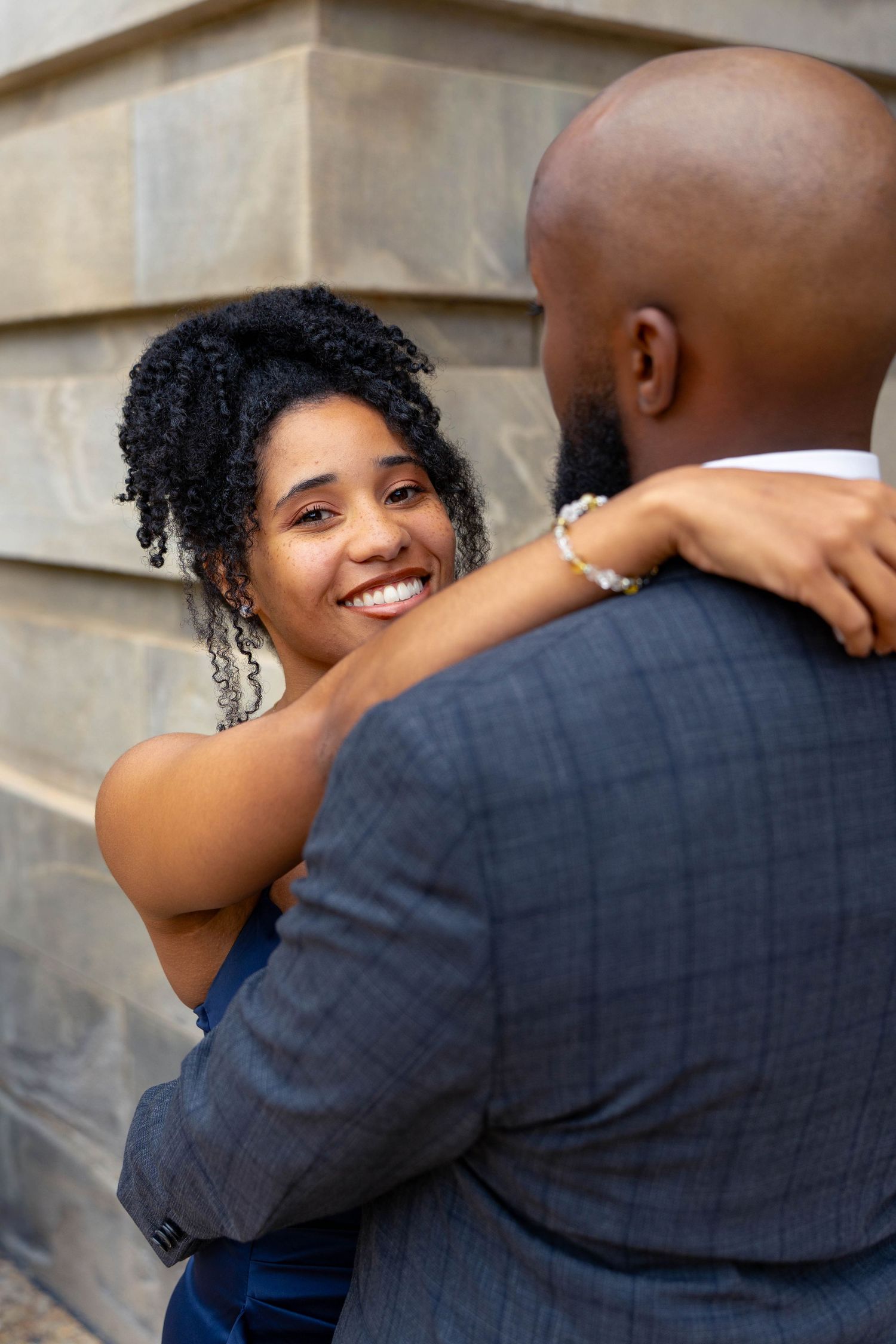 Smiling woman in blue velvet dress embraces man in gray plaid suit outdoors.