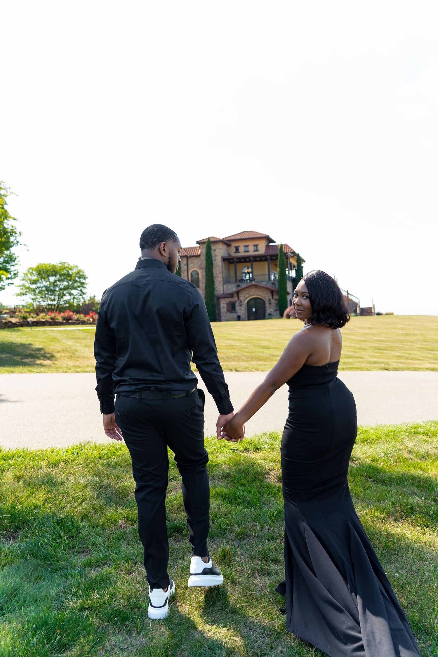 Couple in black formal wear walking hand-in-hand toward elegant stone mansion on sunny day.