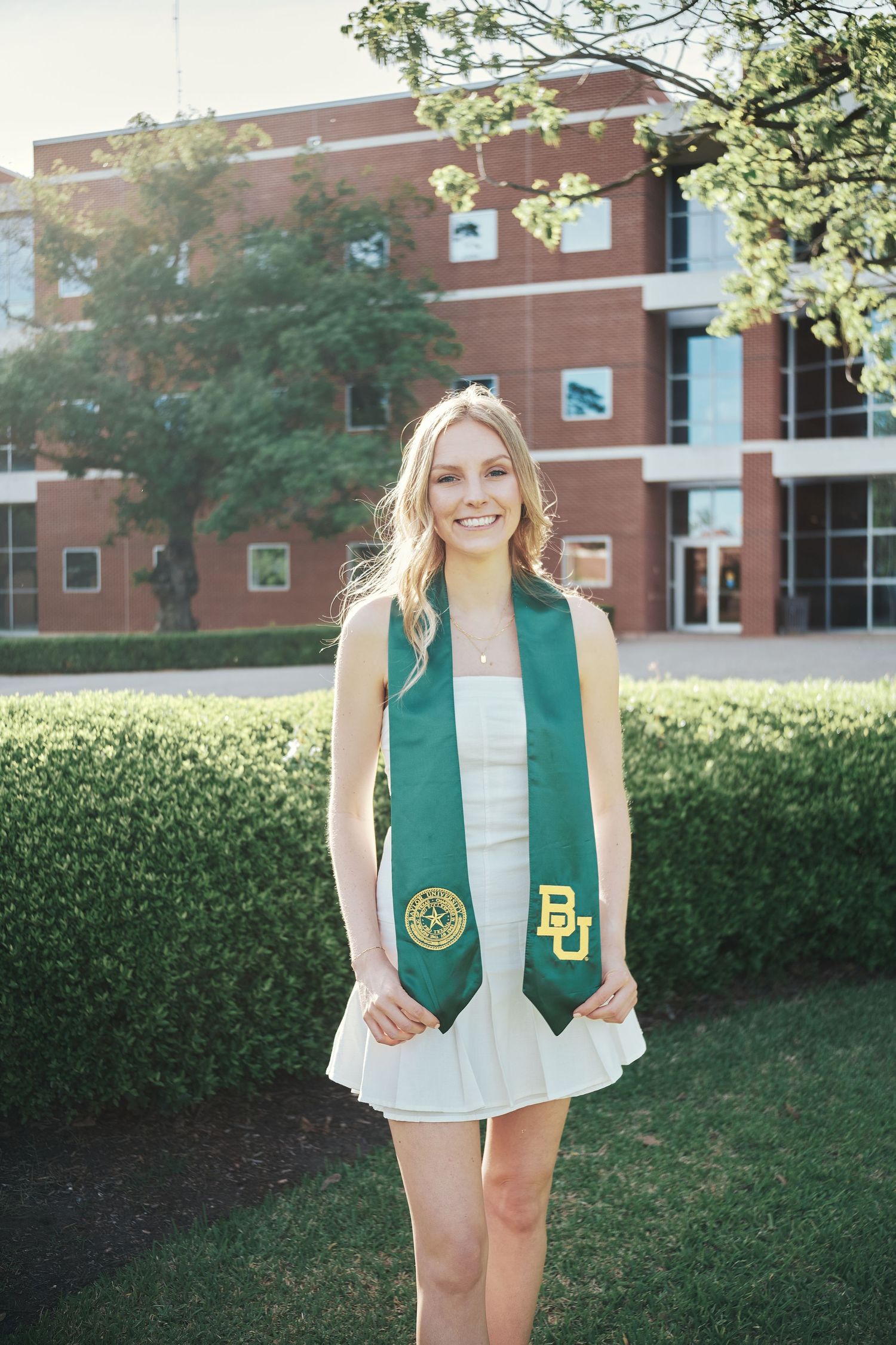 Smiling blonde woman in white dress wearing green Baylor University graduation stole outdoors.