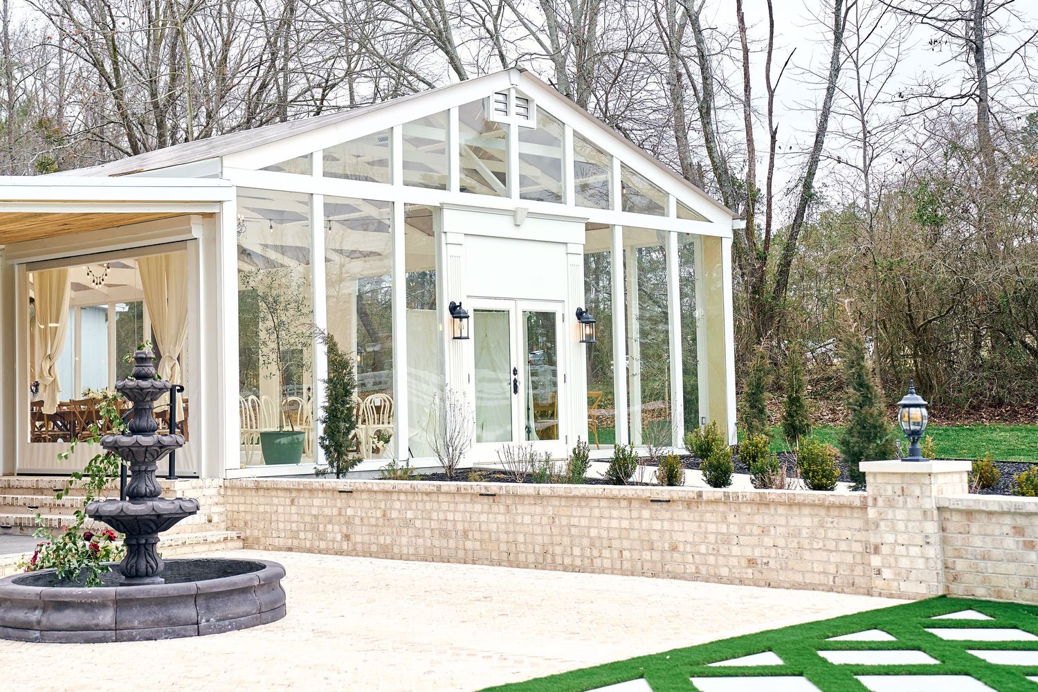 Glass conservatory sunroom with white frame and brick base features an elegant fountain sculpture in the foreground.