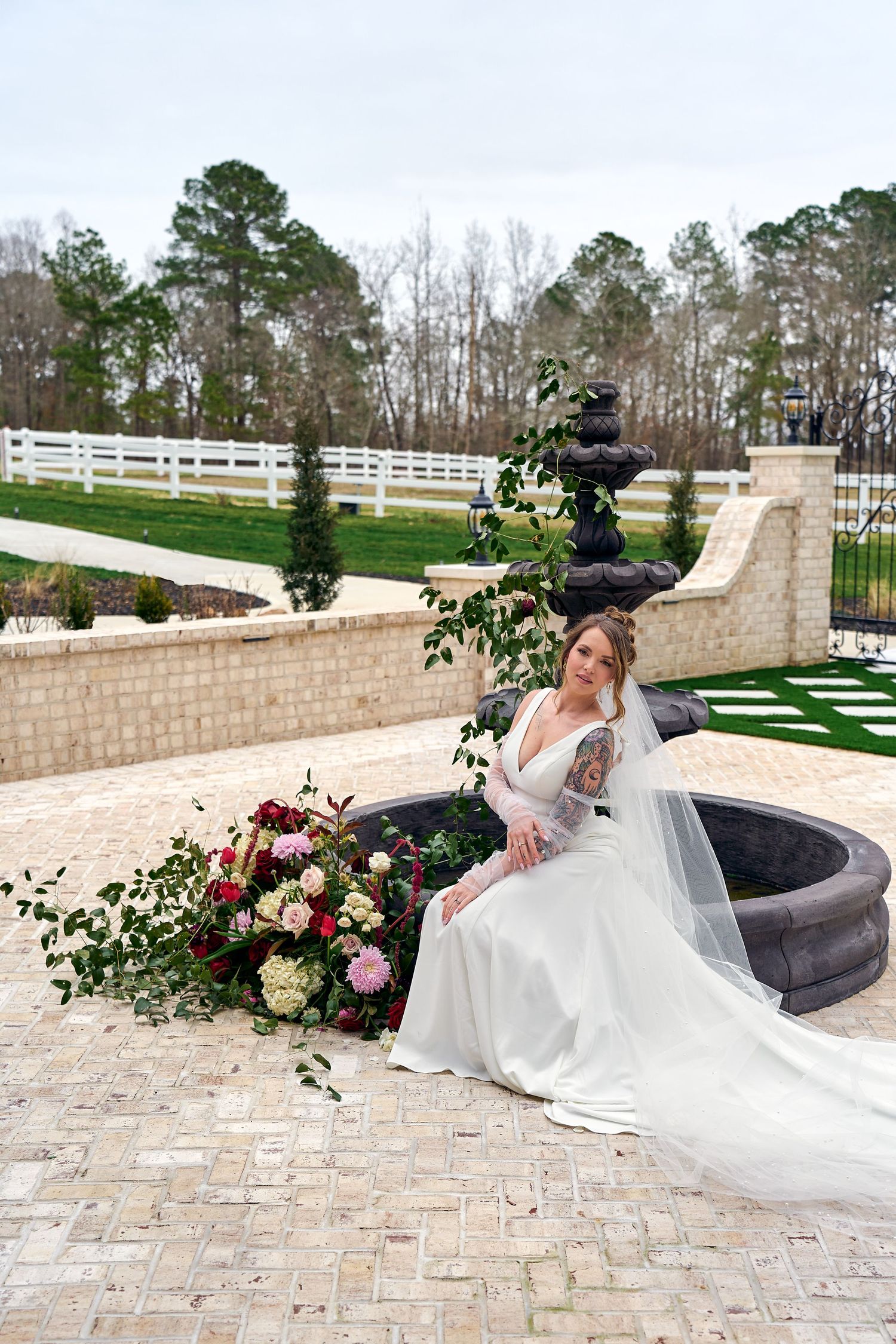 Elegant bridal portrait on stone patio with lush floral arrangement and long flowing veil next to fountain.