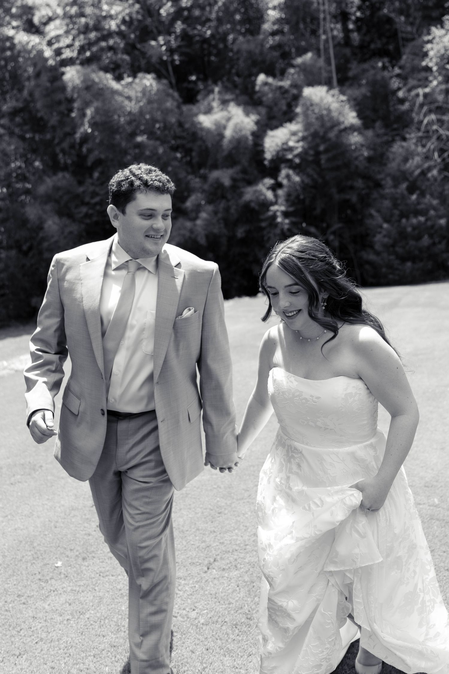 Black and white photo of smiling bride and groom holding hands and walking together on their wedding day.