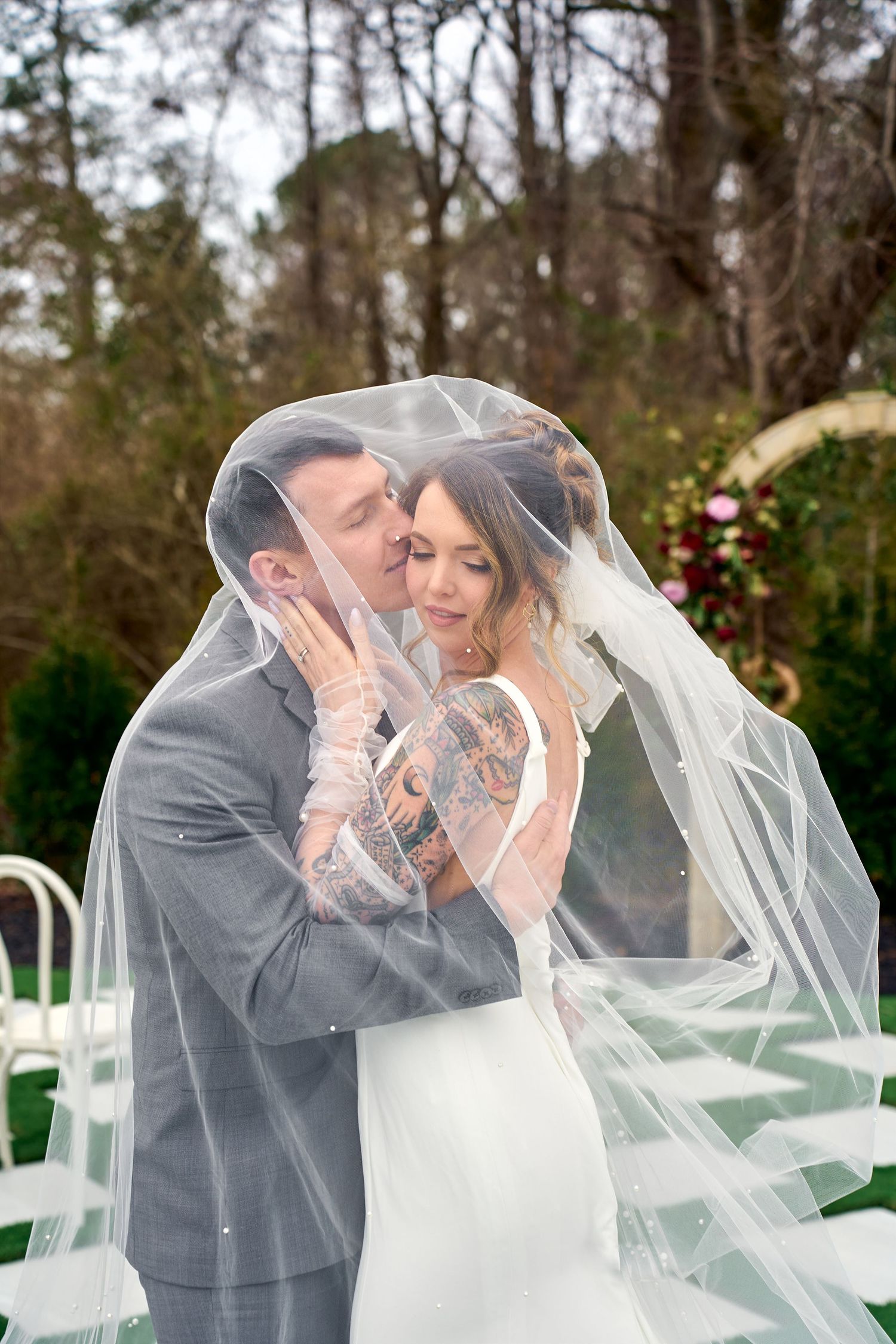 A romantic wedding moment captured outdoors with a bride in white gown and flowing veil embracing in gray suit against garden backdrop.
