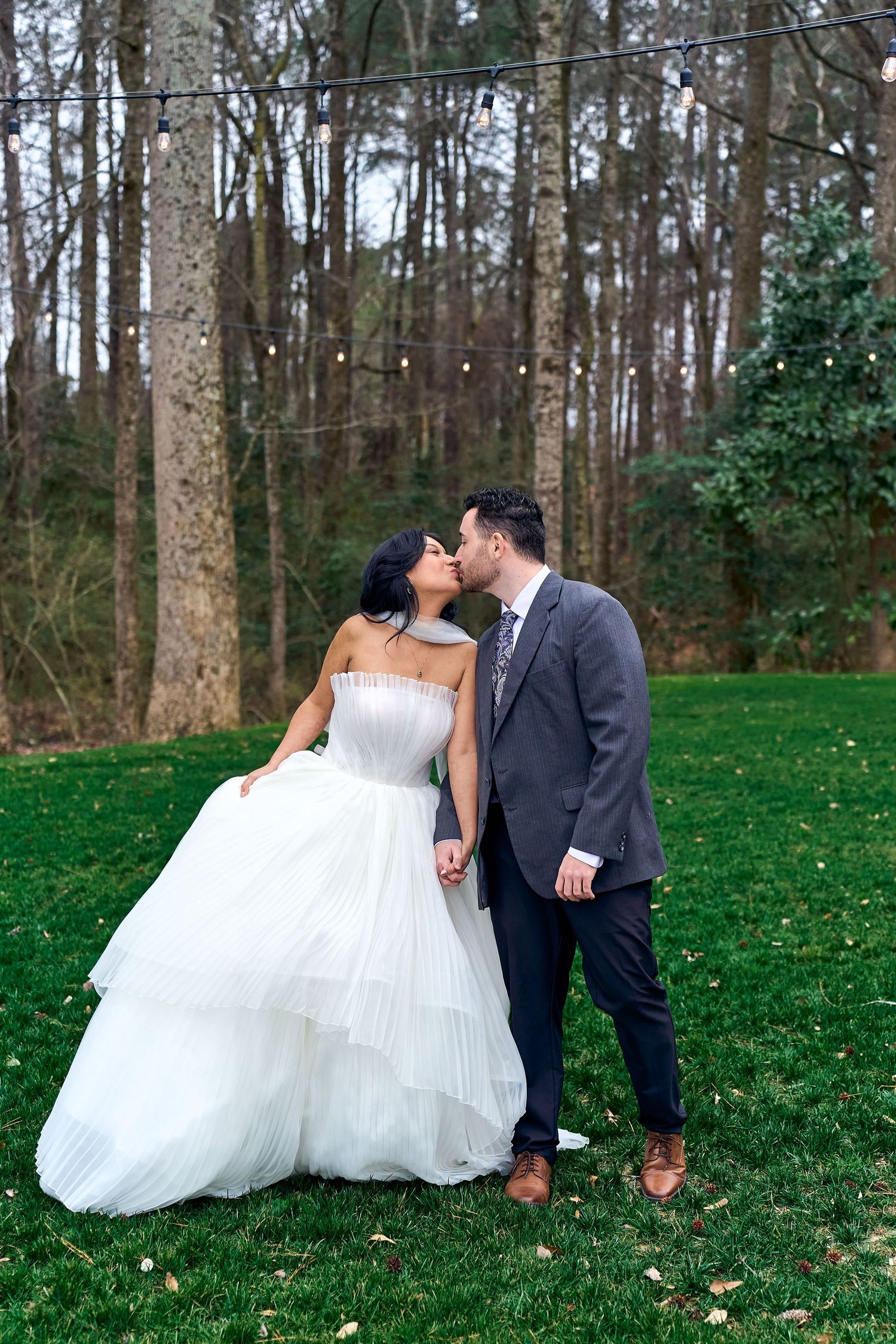 A couple shares a romantic kiss on their wedding day in a wooded outdoor setting with tall trees and green grass.