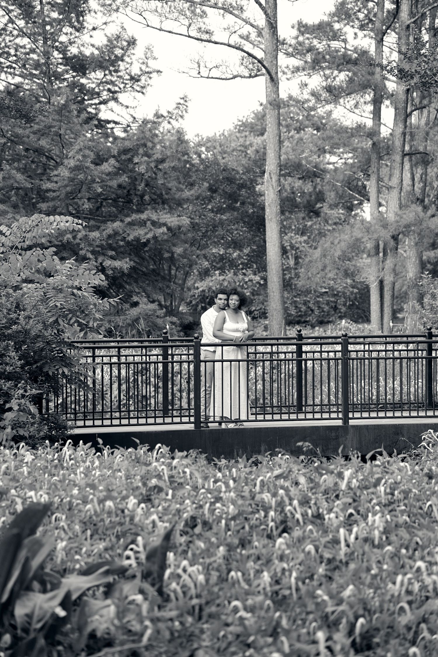 A couple standing on a deck surrounded by lush gardens and flowering plants in black and white.