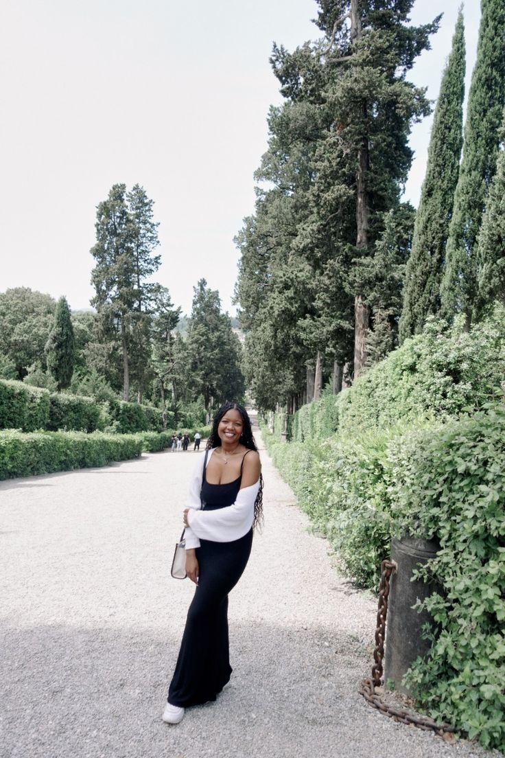 Smiling woman in black outfit with white wrap strolling through a lush green garden pathway.