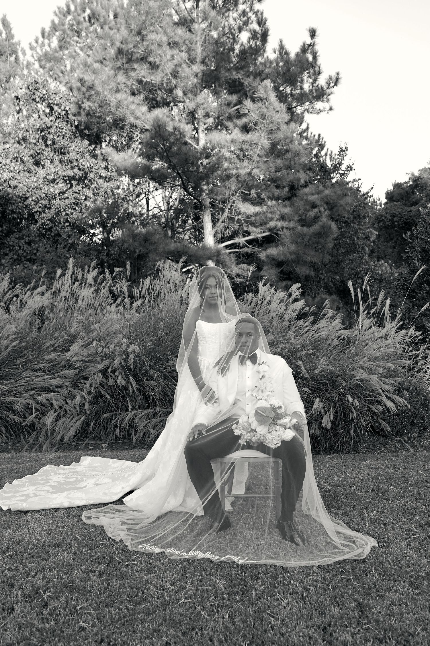 Bride and groom posing in garden, bride's long veil draped over seated groom outdoors.