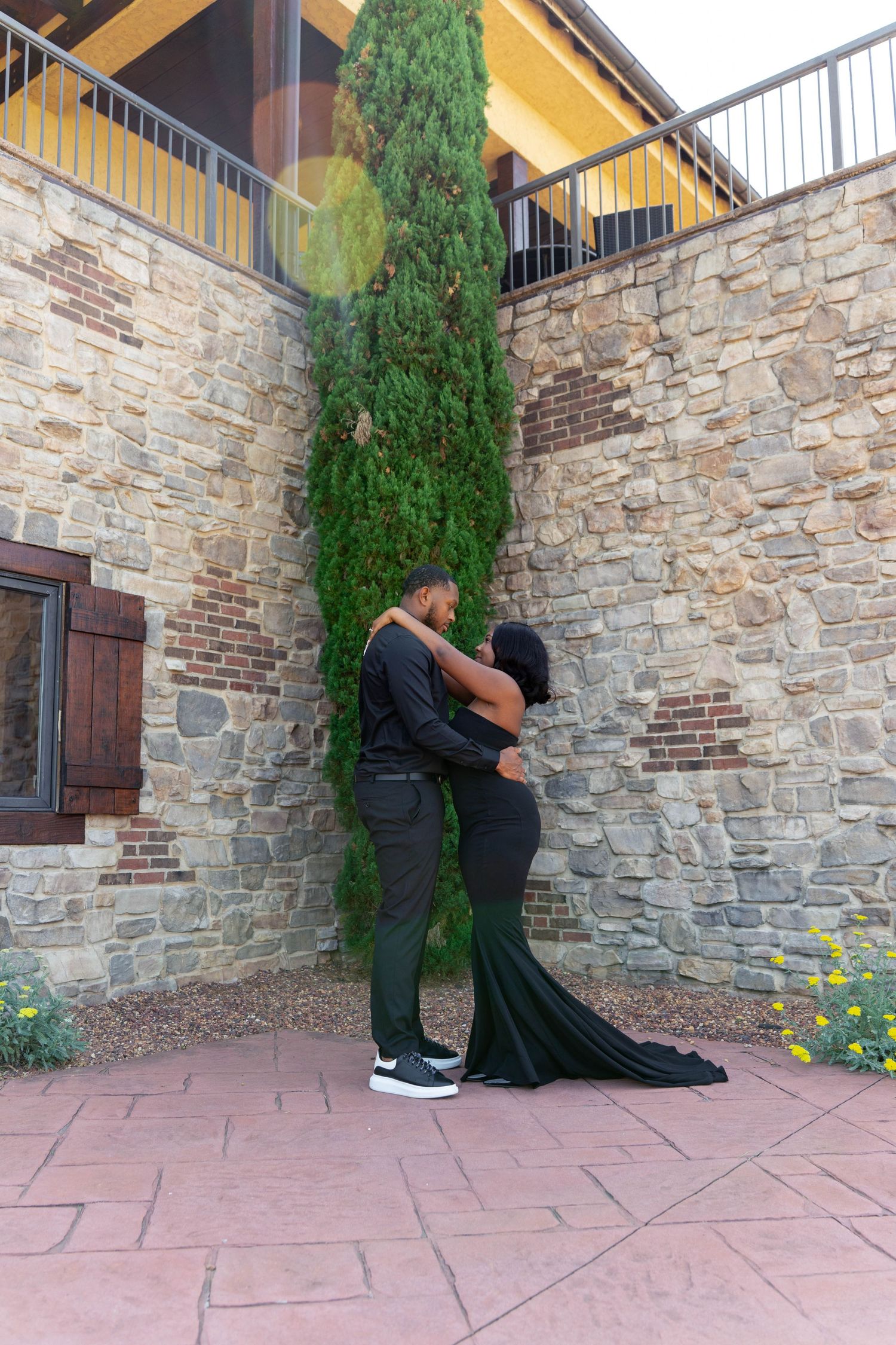 Couple in all-black outfits sharing a kiss beside tall cypress tree against rustic stone wall backdrop.