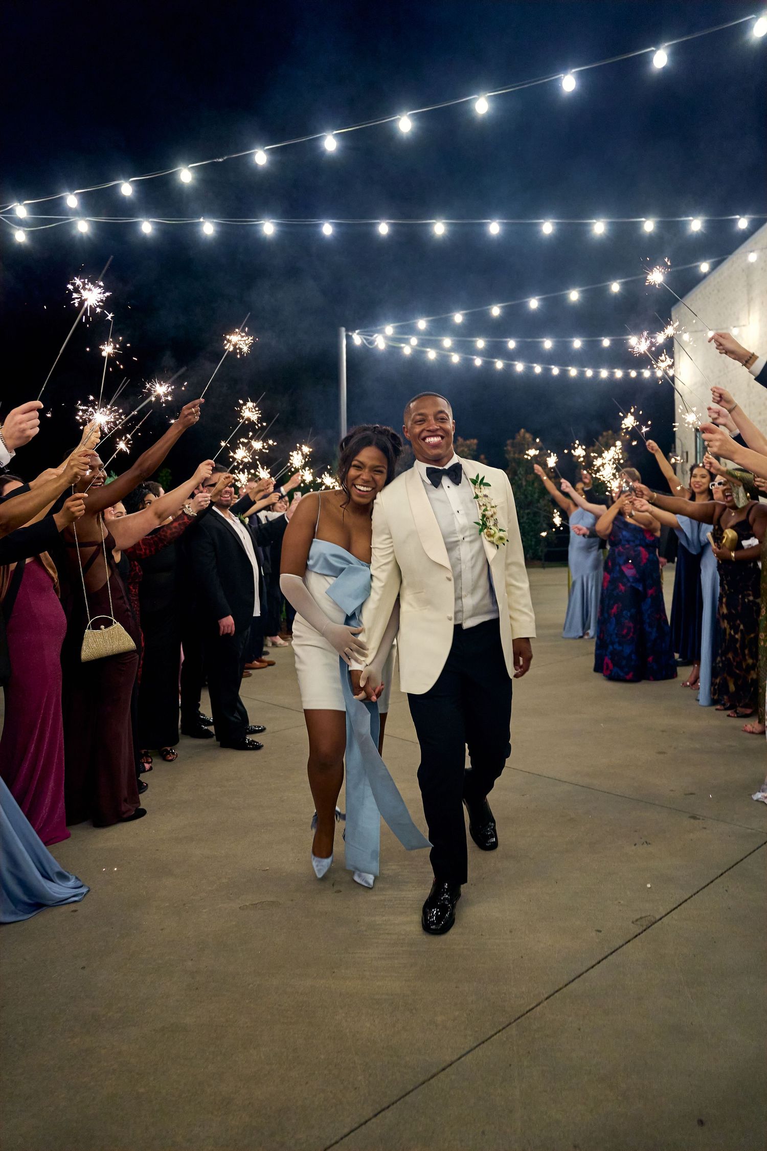 Newlyweds walk through a sparkler send-off tunnel under string lights at their outdoor wedding exit.